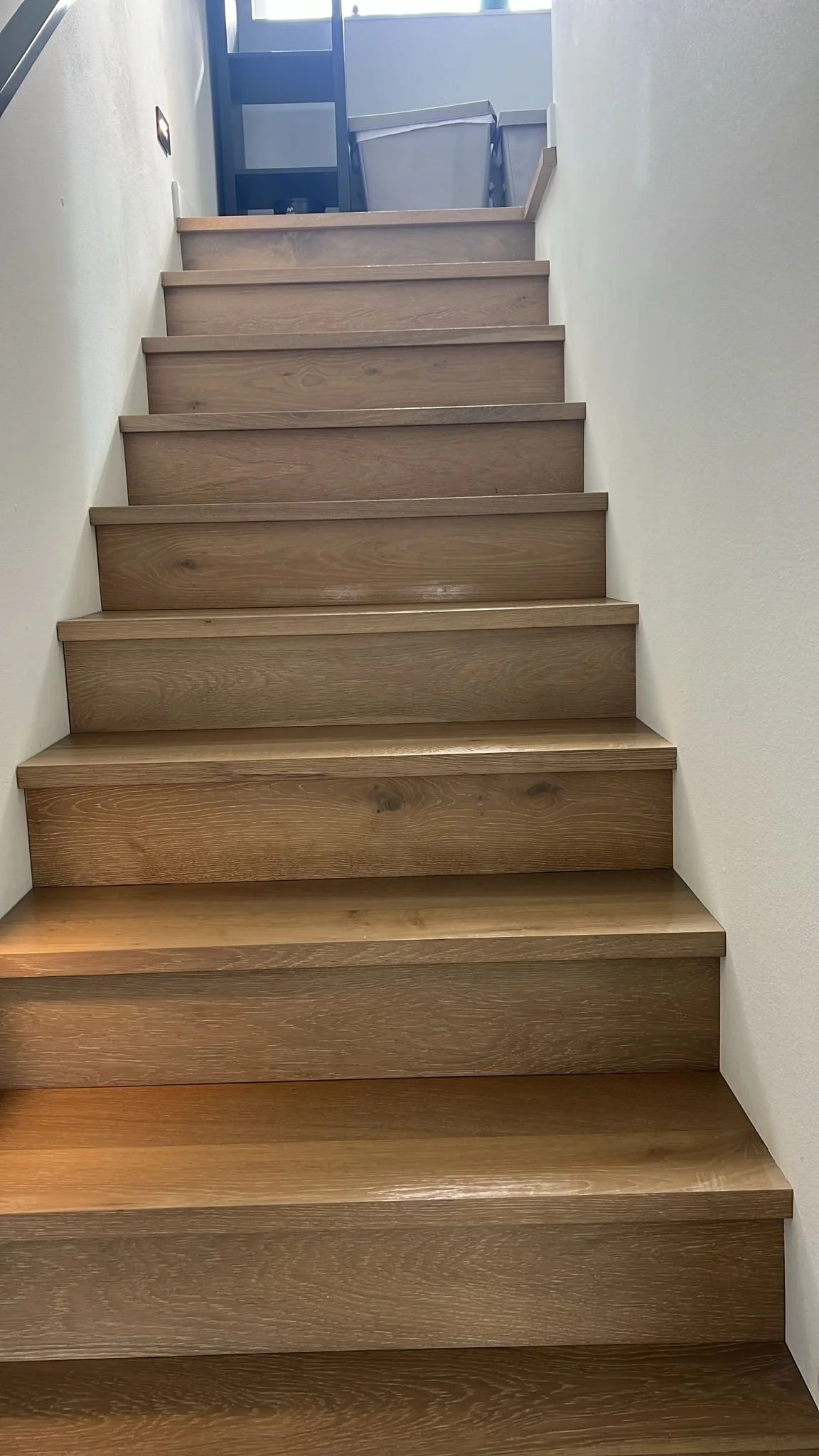 Wooden staircase ascending with a wall on each side, sunlight coming from a window at the top.