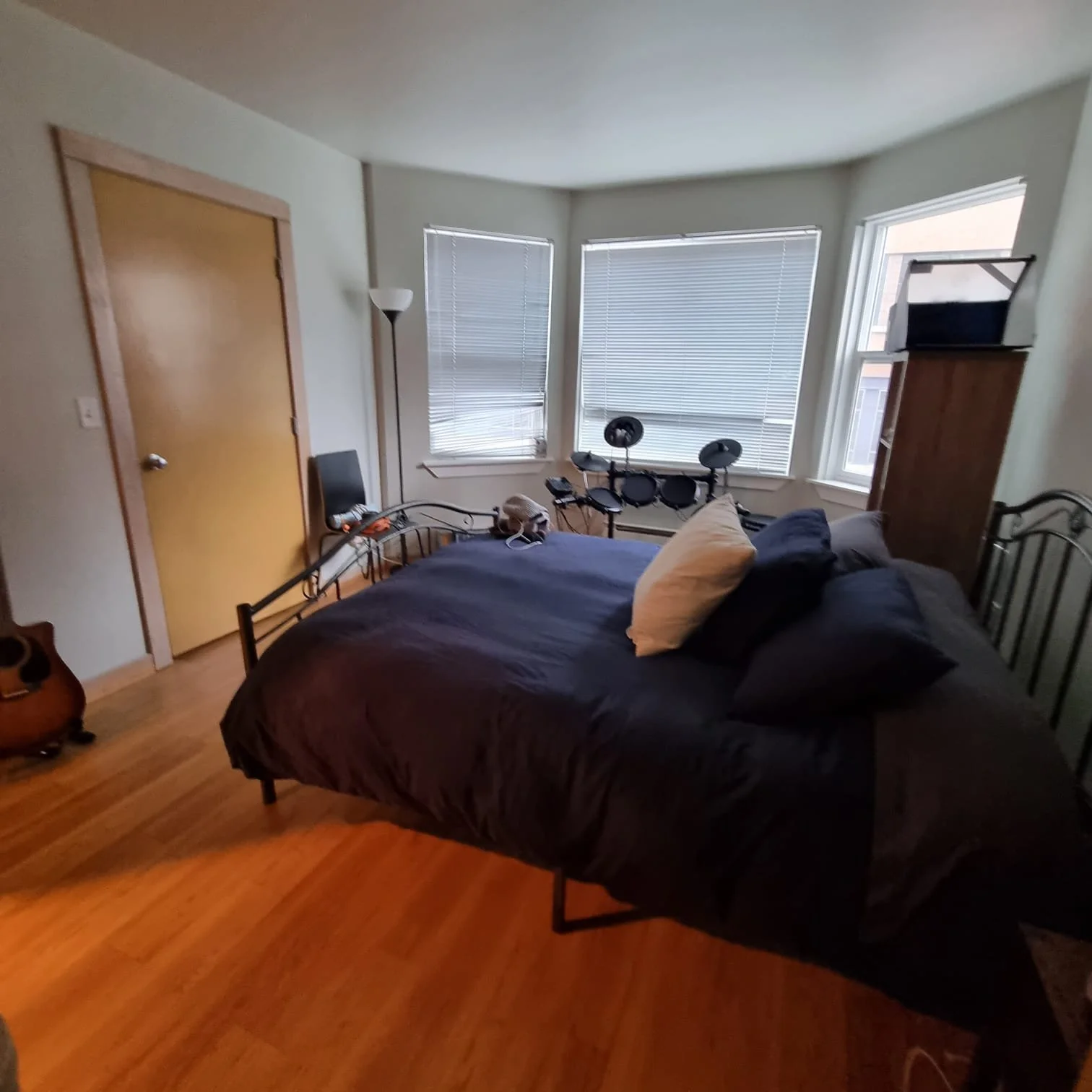 A bedroom with a bed covered with a dark duvet and pillows, a guitar on the floor, a chair, a tall lamp, three windows with blinds, and an electronic drum set near the windows.