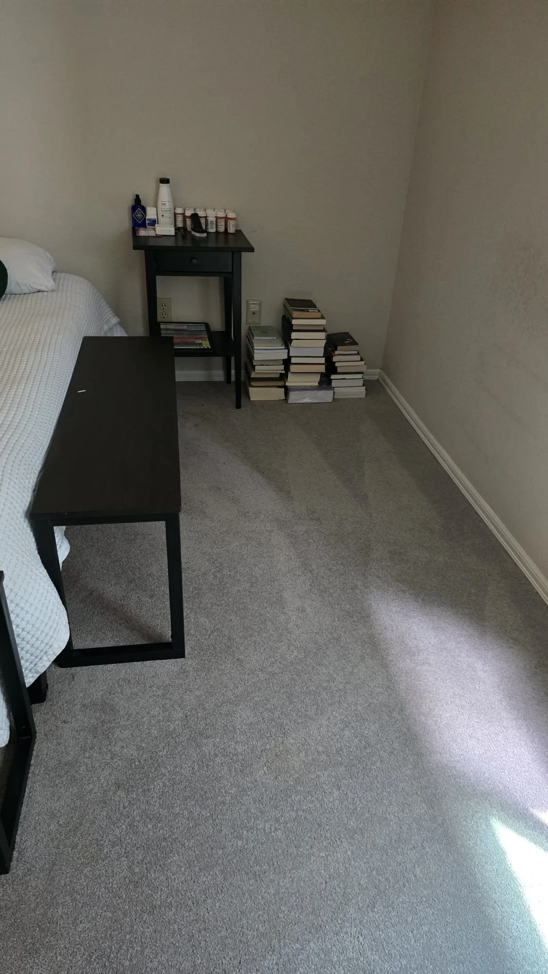 A corner of a bedroom with beige carpet, a bed with white bedding, a black nightstand with miscellaneous items on top, and a pile of books stacked against the wall.