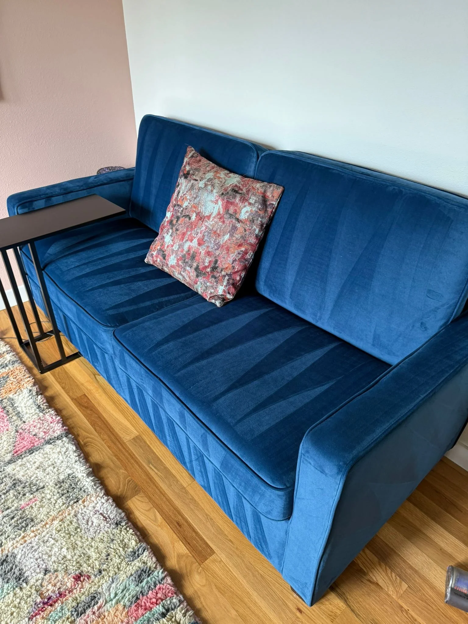 Blue velvet sofa with a patterned throw pillow, situated next to a multicolored shag rug and a small black side table in a room with hardwood flooring and two-toned walls.