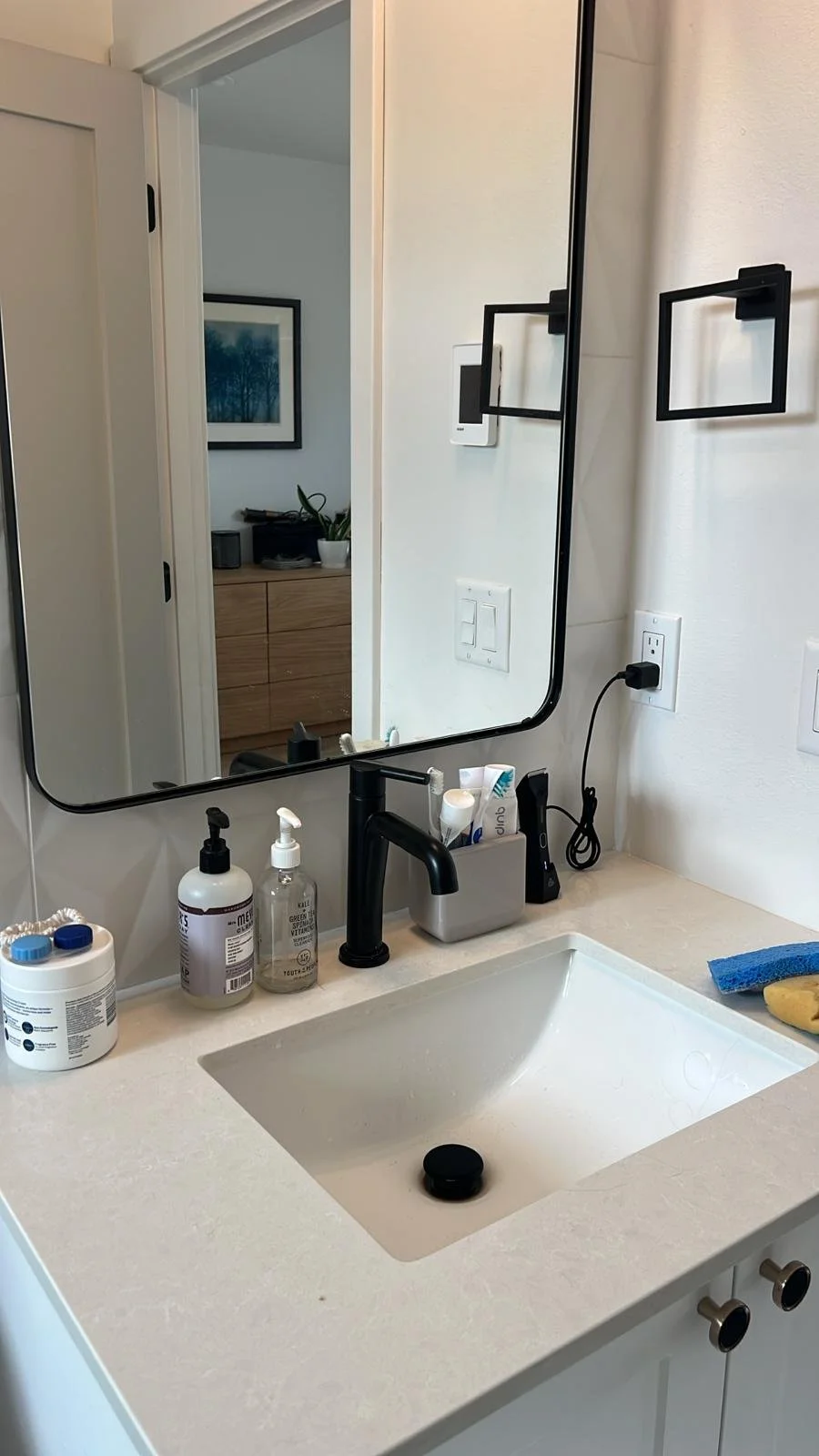 Bathroom vanity with a mirror, black faucet, containers of hand soap and lotion, toothbrush holder, sponge, and a towel.