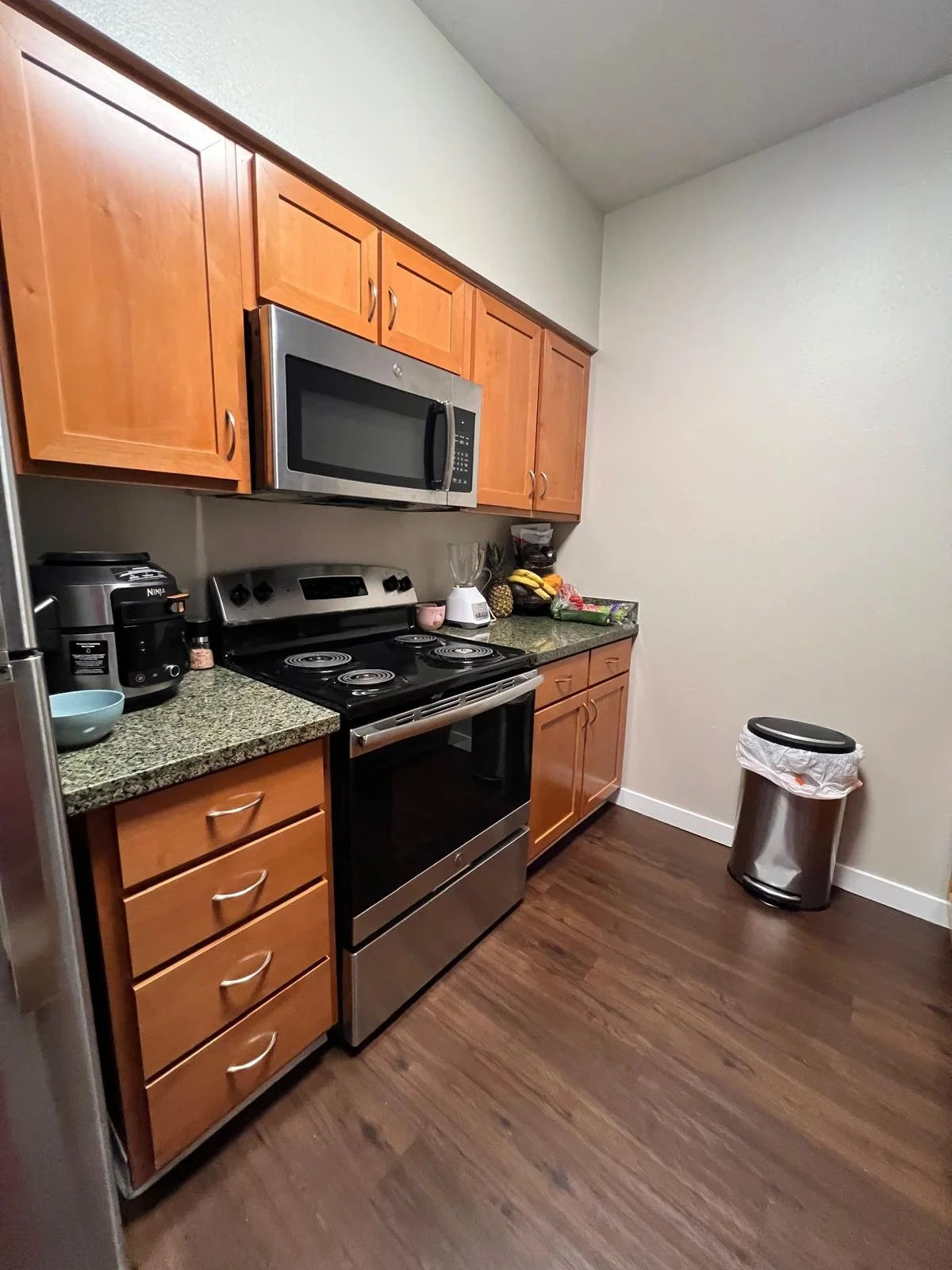 Kitchen with wooden cabinets, stainless steel stove and microwave, granite countertop, small appliances, and a trash bin.
