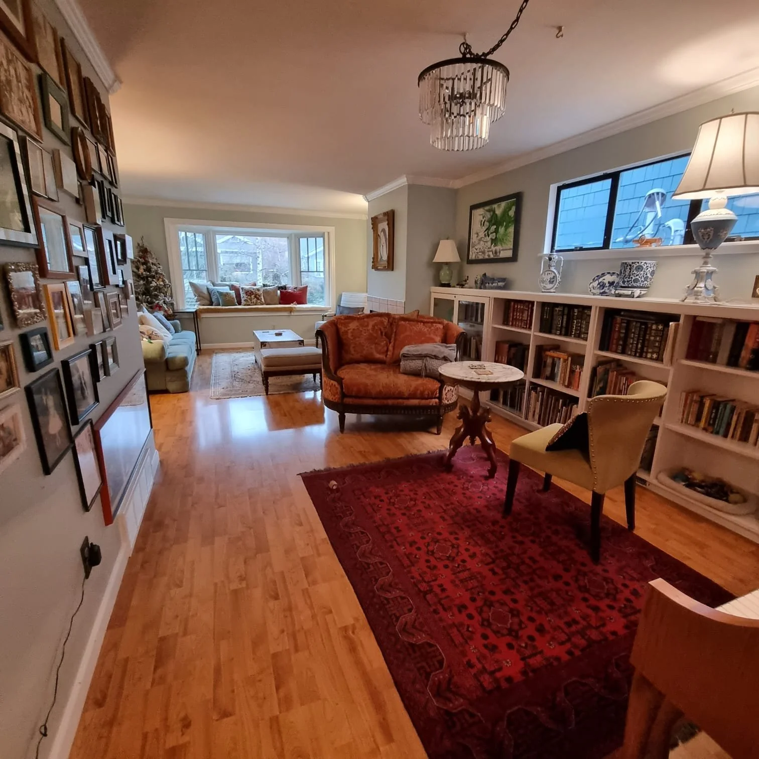 A cozy living room with a bay window seat filled with pillows, a vintage sofa, bookshelves with decorative items, and hardwood floors with an area rug. There is a chandelier hanging from the ceiling and various lamps providing warm lighting.