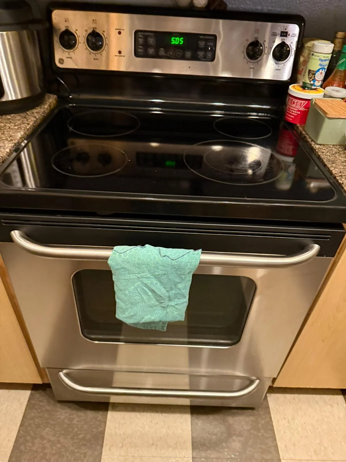 Stainless steel electric stove and oven with four burners, a green towel hanging on the oven handle, some kitchen items like seasoning bottles, a container of ice cream, and a box on the countertop.
