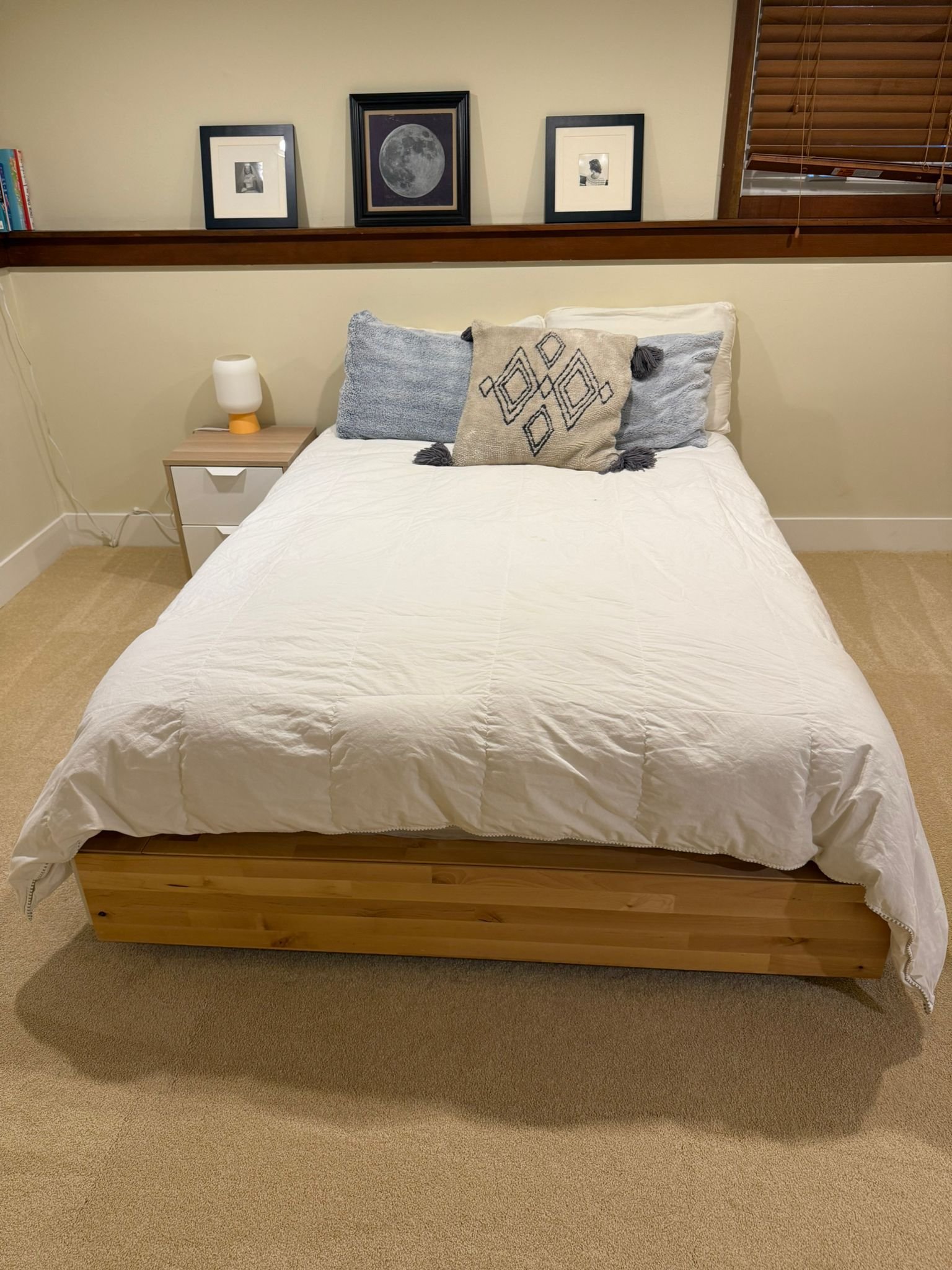 A neatly made bedroom bed with white bedding, two gray pillows, and a decorative throw pillow with black geometric patterns. A small wooden nightstand with a white lamp on it is beside the bed. Above the bed, there are three framed photos, including 