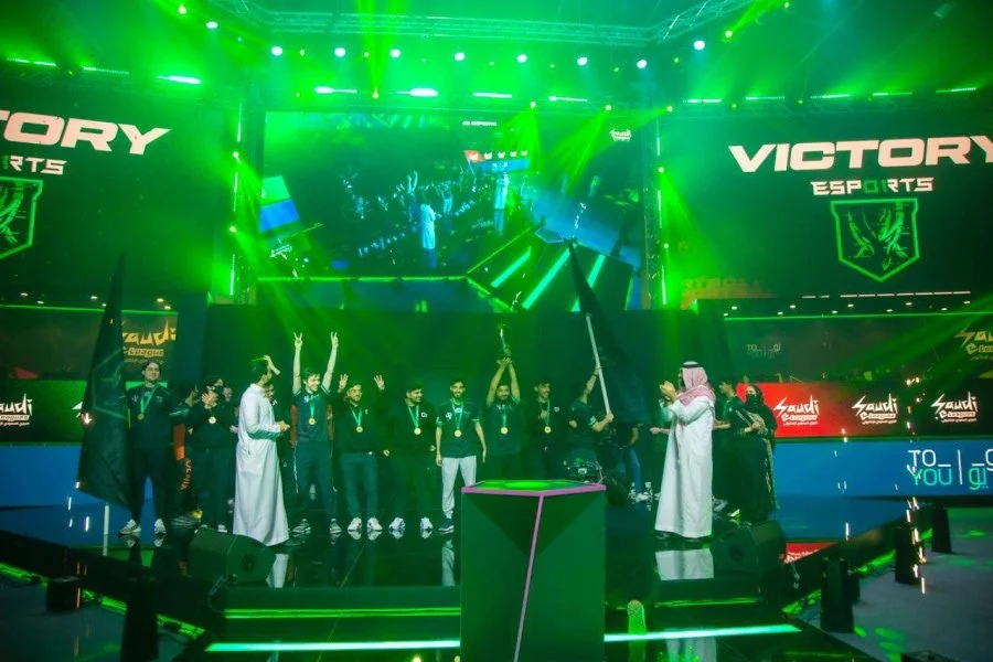 ESports team on stage after winning, holding trophy, surrounded by fans and moderators in a green-lit arena with large screens displaying victory and team logos.