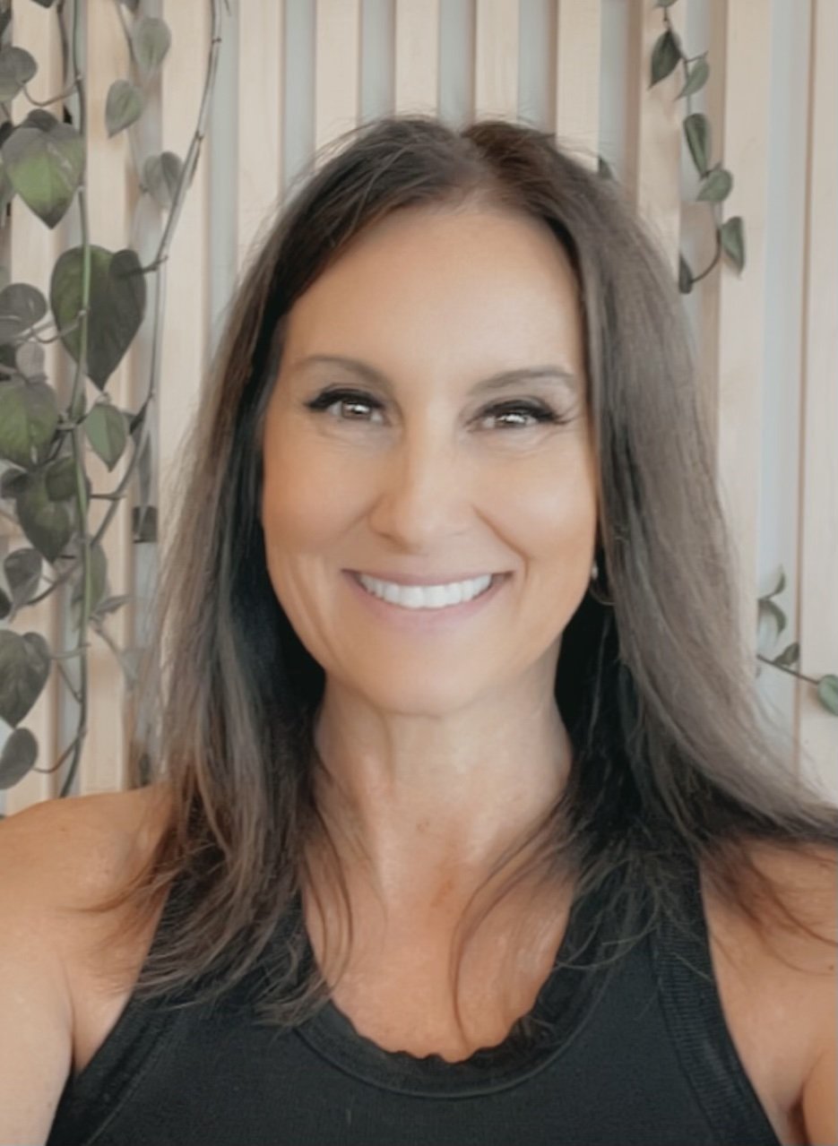A woman smiling with straight brown hair, wearing a black top, standing in front of a wooden fence with green leafy vines.