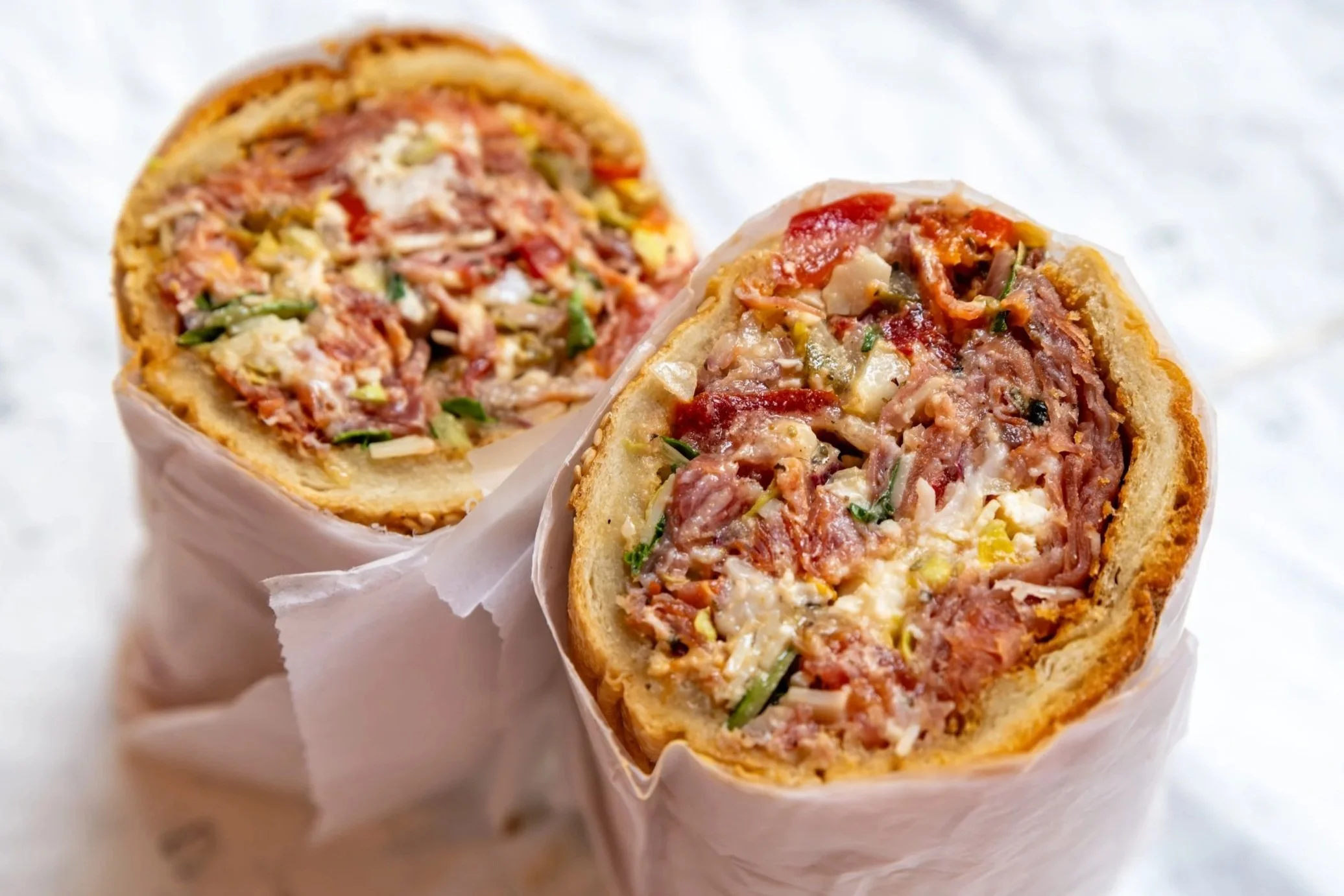 Close-up of two halves of a savory meat and vegetable stuffed bread roll wrapped in parchment paper on a white surface.