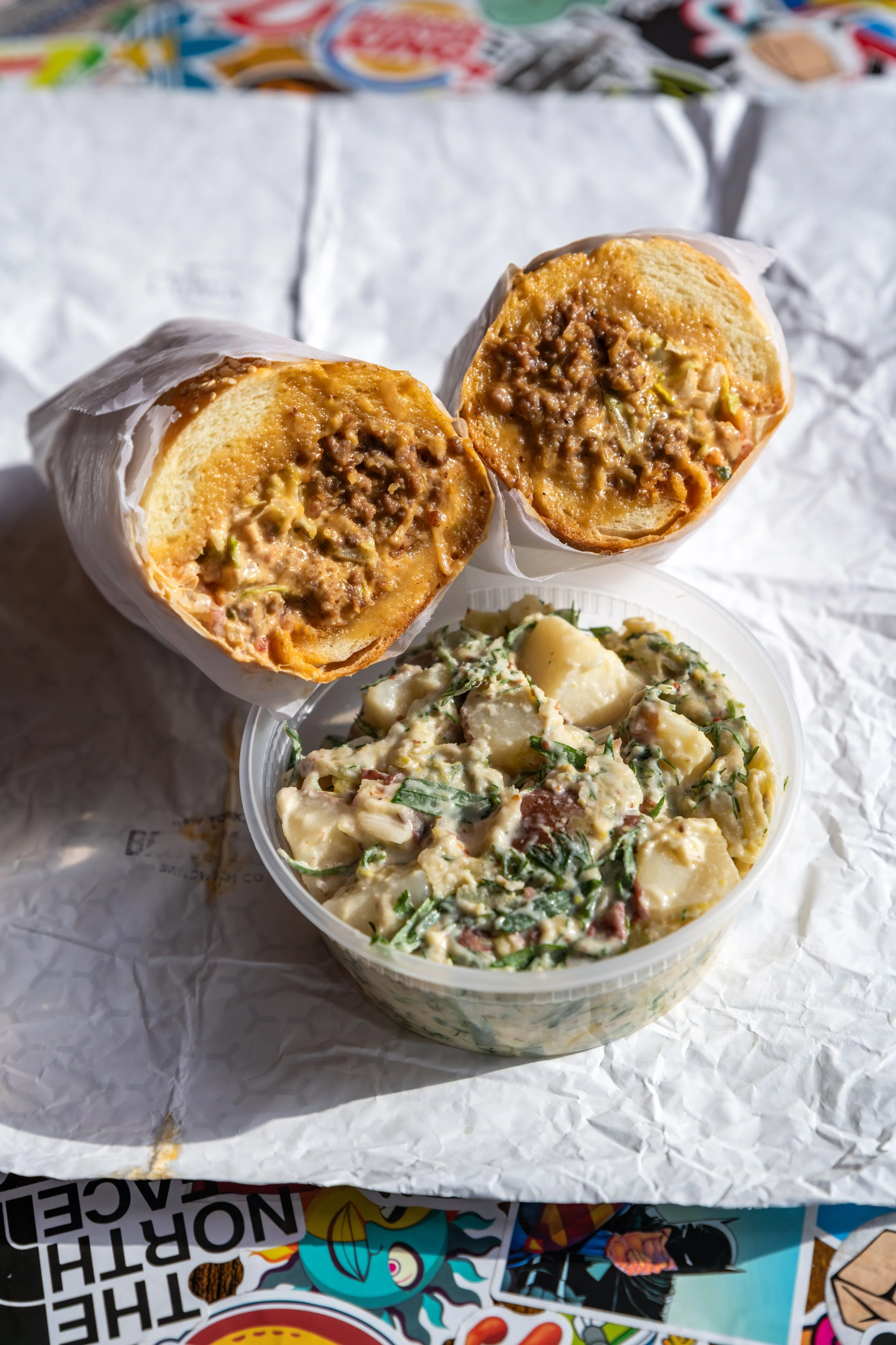 A sandwich with seasoned beef, cheese, and vegetables, cut in half, resting on wax paper. Next to it is a container of potato salad with potatoes, bacon, green onions, and dressing.