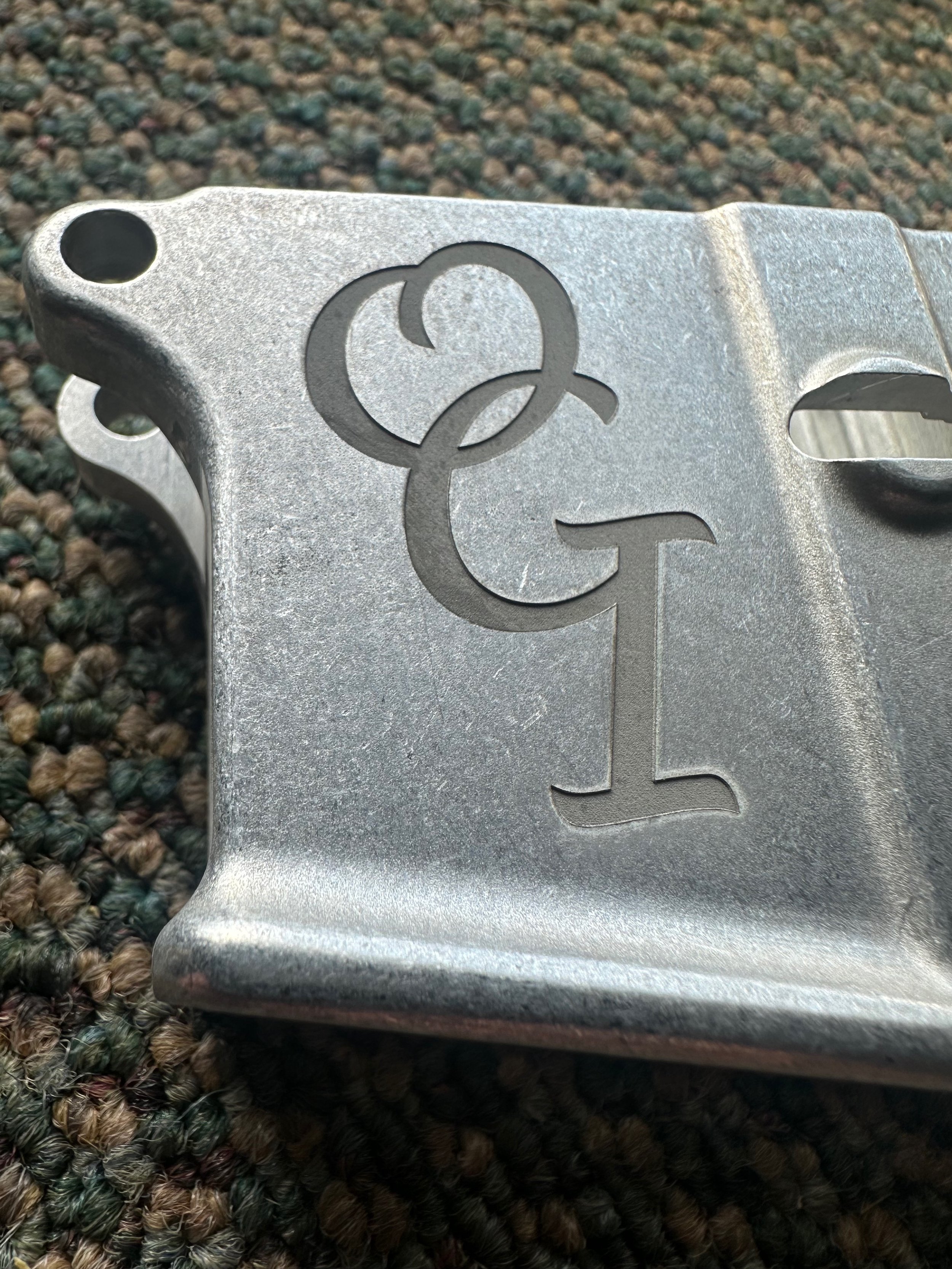 Close-up of a metallic object with laser-etched letters "CGI" on its surface, placed on multicolored, textured carpet.