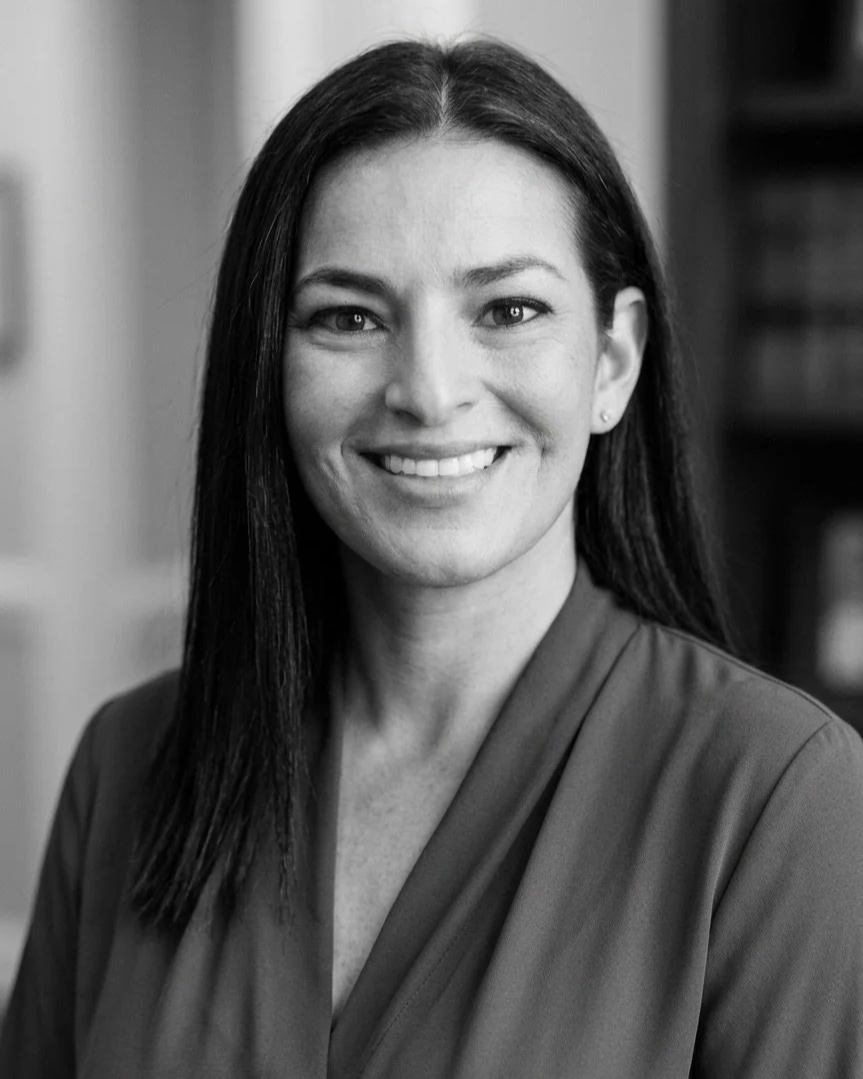 Black and white portrait of a smiling woman with long dark hair, wearing a dark blouse, positioned in front of a blurred background.