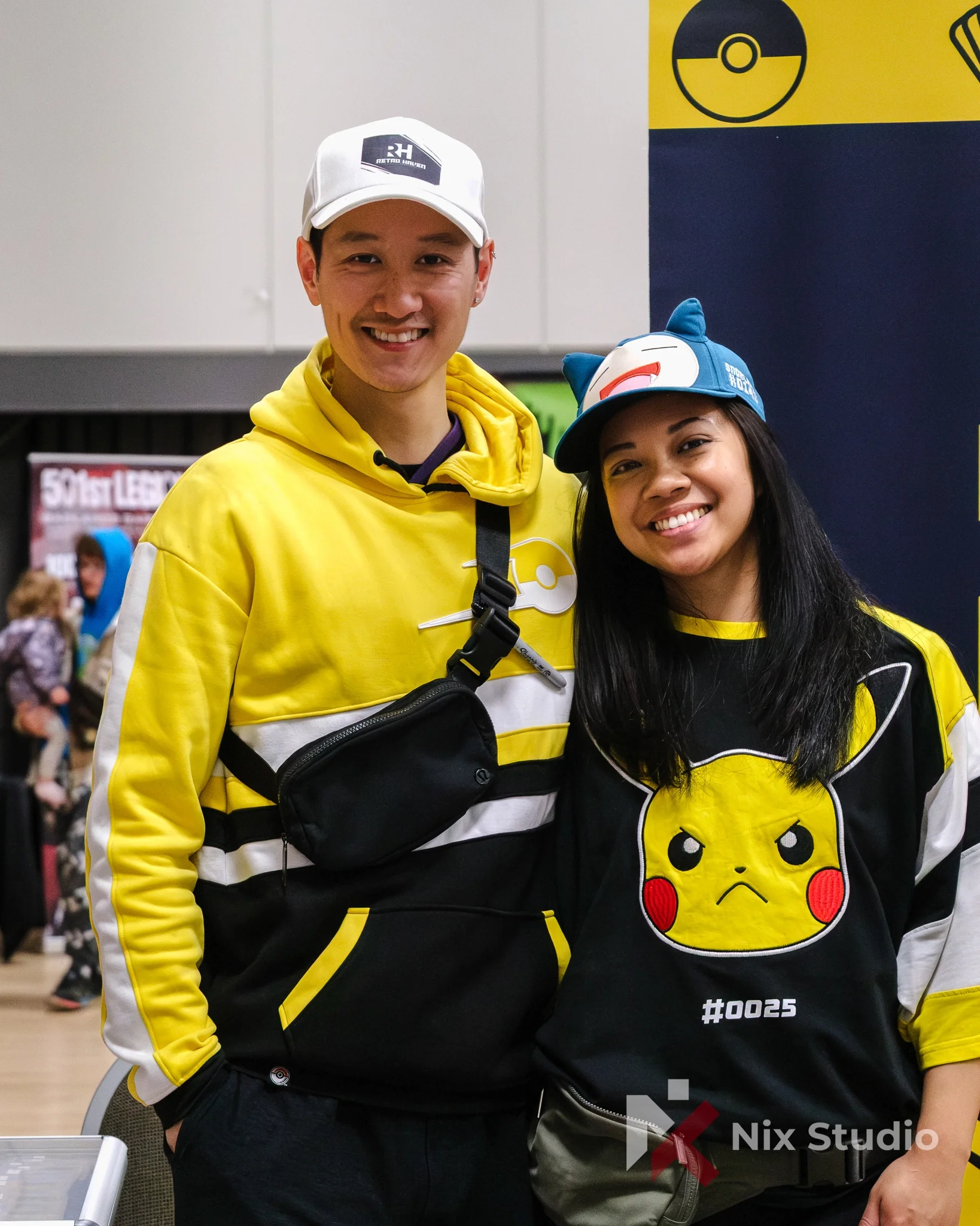 Two young people at an event, both wearing yellow and black Pikachu-themed hoodies and black shirts with Pikachu graphics. They are smiling and posing for the camera. The man is wearing a white cap with a black patch that says 'RETRO HAVEN,' and the 