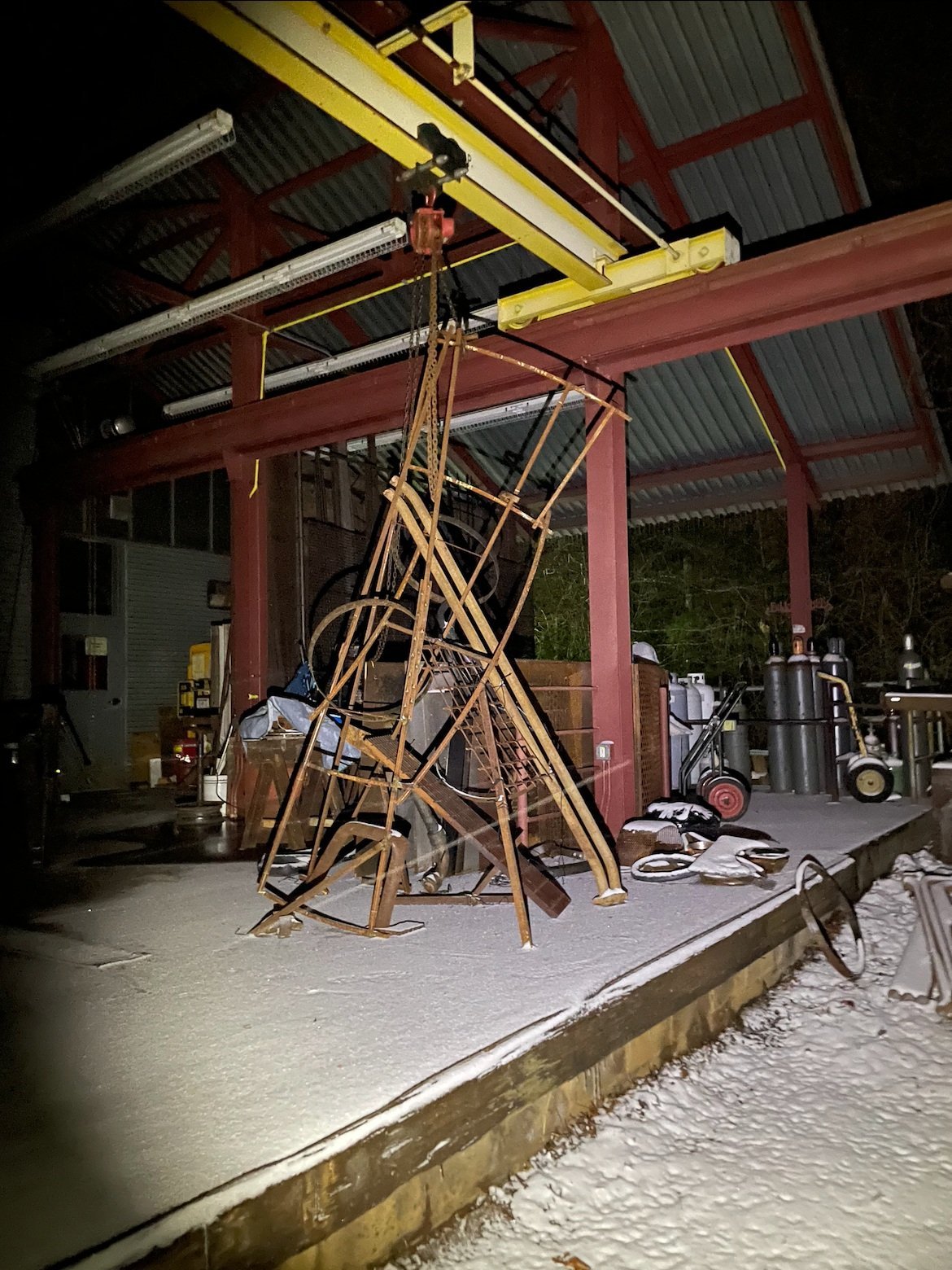 Outdoor industrial area with metal frames, a hoist system overhead, various pipes and equipment, and a light dusting of snow.