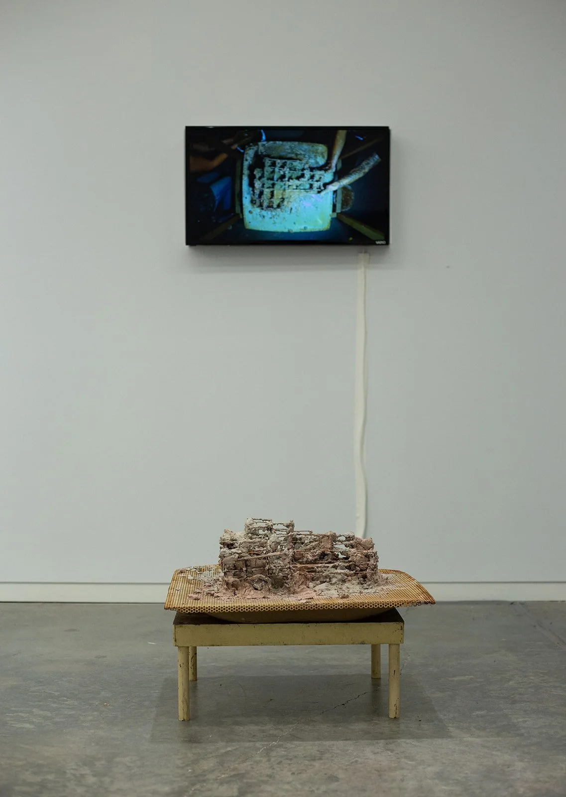 Art installation with video screen and clay sculpture on small table in gallery space.