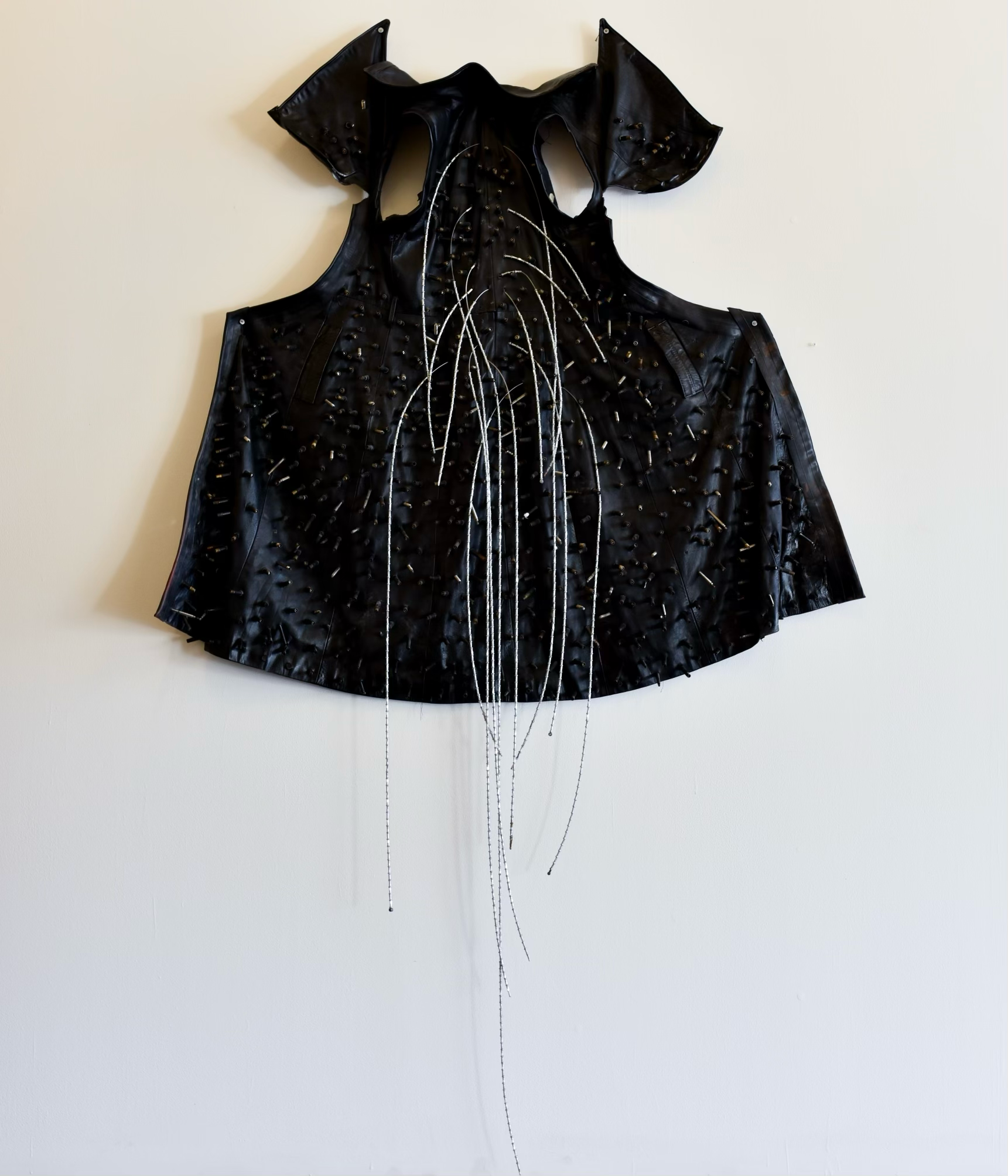 Black leather vest with metal spikes and decorative metal chains hanging down, displayed on a white wall.