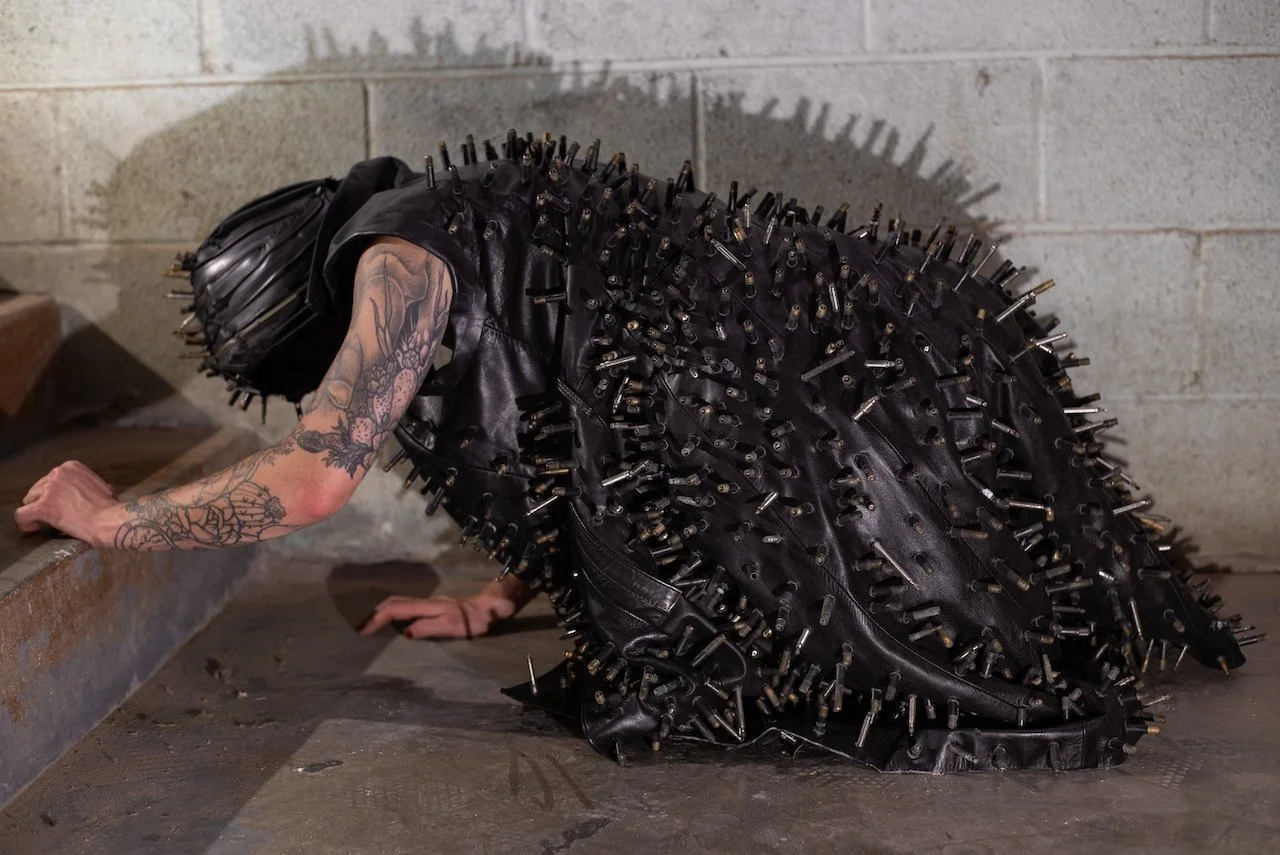 A person wearing a bulky leather outfit covered in metal spikes is crouched on the floor in an industrial setting with tattoos visible on their arm.