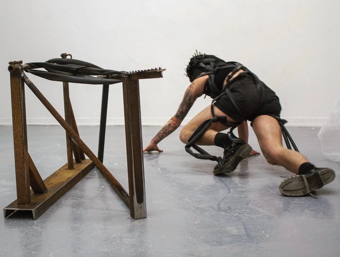 Person wearing a dark, strapped outfit and crawling on a floor in a gallery-like setting near an abstract metal sculpture.
