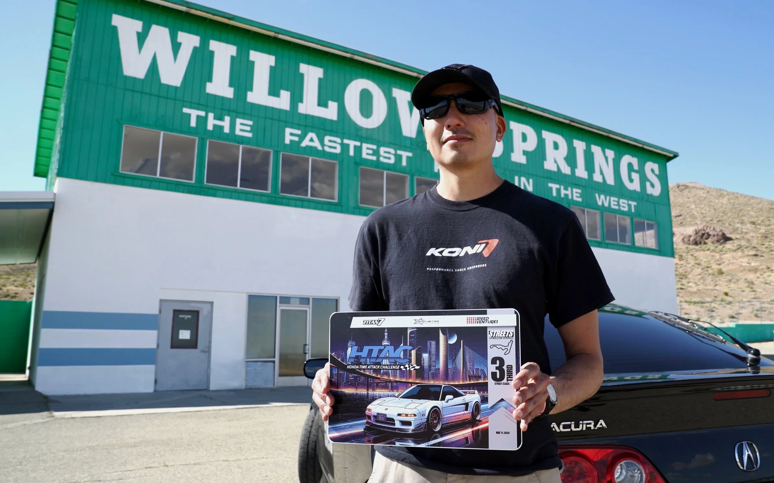A man wearing sunglasses and a black t-shirt stands outside holding a poster of a white sports car with a city skyline background. Behind him is a green building with large white letters that say 'WILLOW SPRINGS' and 'THE FASTEST IN THE WEST'.