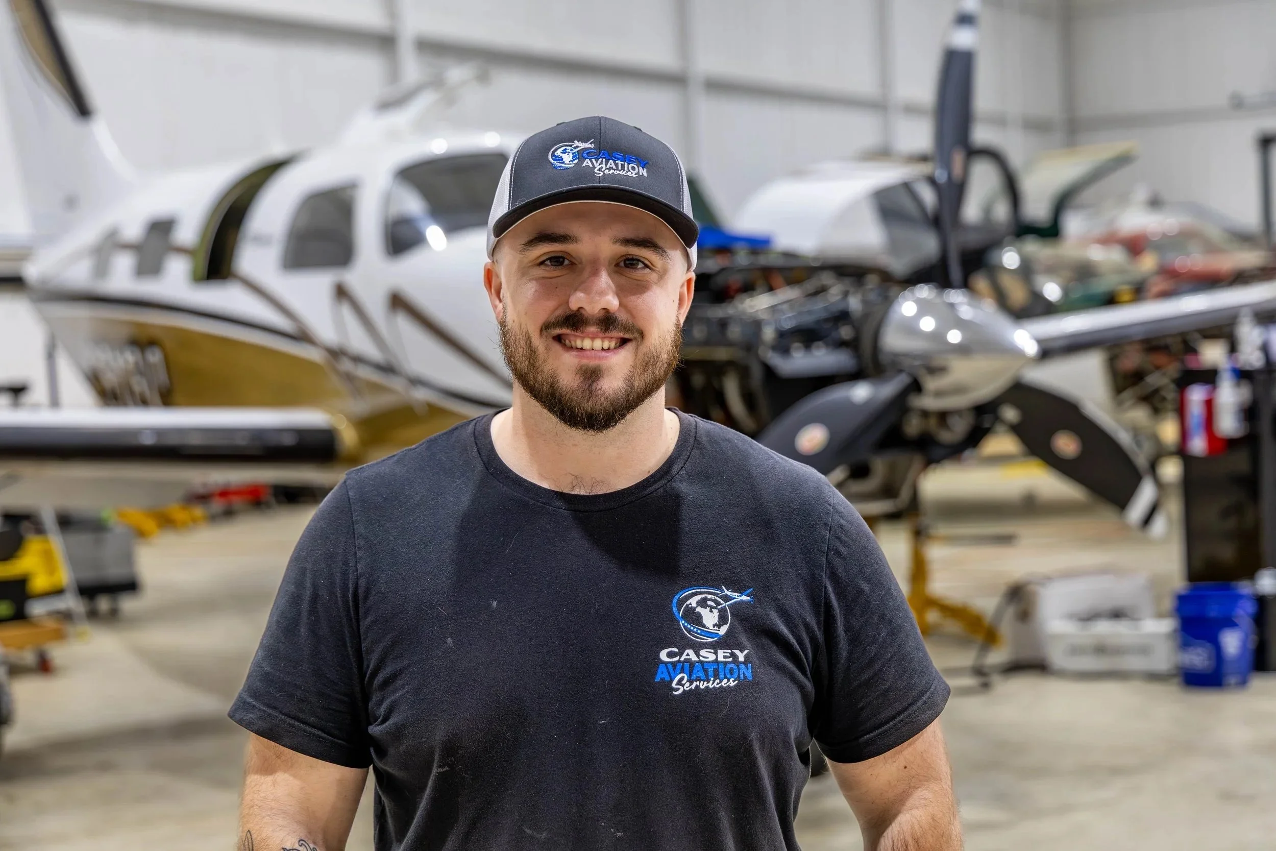 A smiling man wearing a black Casey Aviation Services t-shirt and cap, standing in an aircraft workshop, with a small airplane in the background.