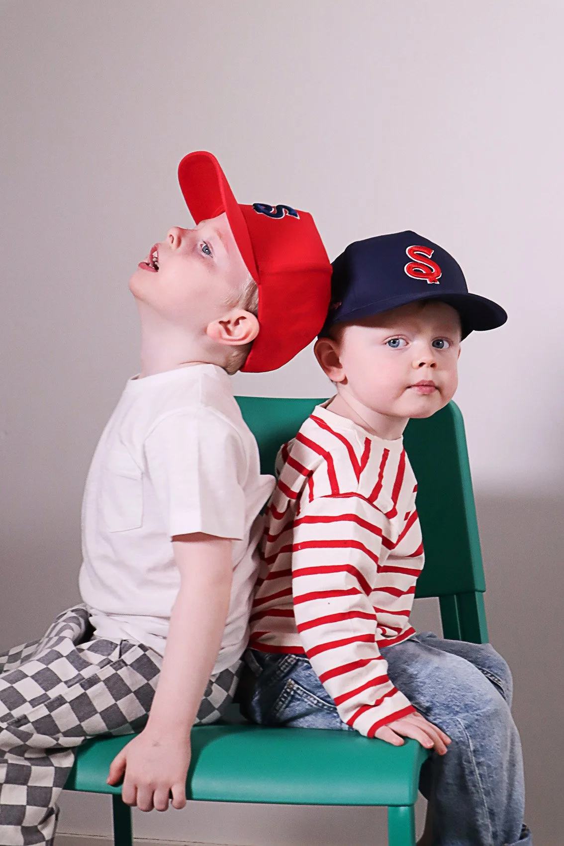 Two young children sitting back-to-back on a green chair, both wearing baseball caps; one in a red cap with a white shirt and checkered pants, and the other in a blue cap with a red and white striped shirt and jeans.