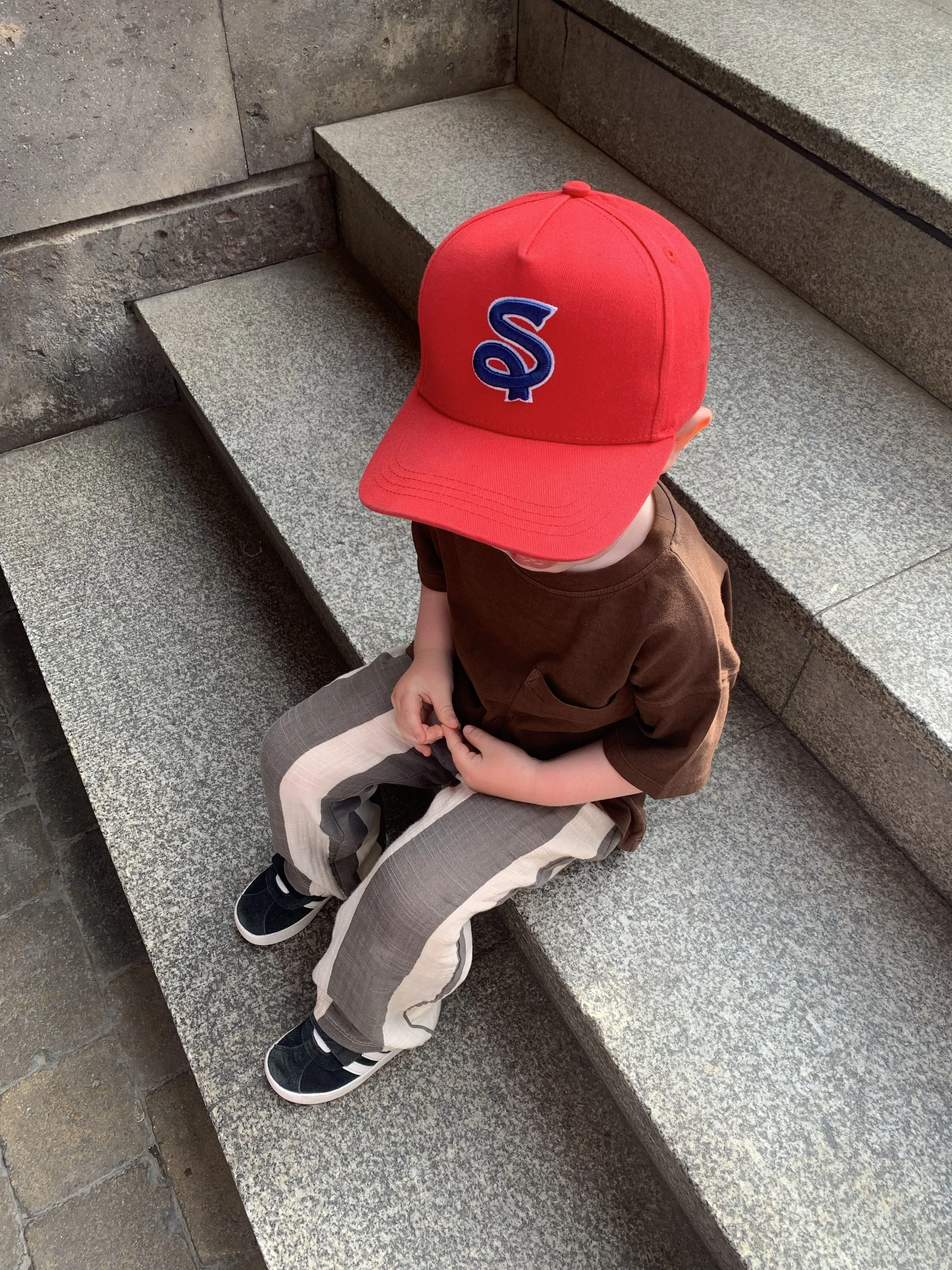 A young child sitting on concrete steps, wearing a red Smallz logo baseball cap with a blue and white logo, a brown shirt, beige and brown striped pants, and black sneakers.