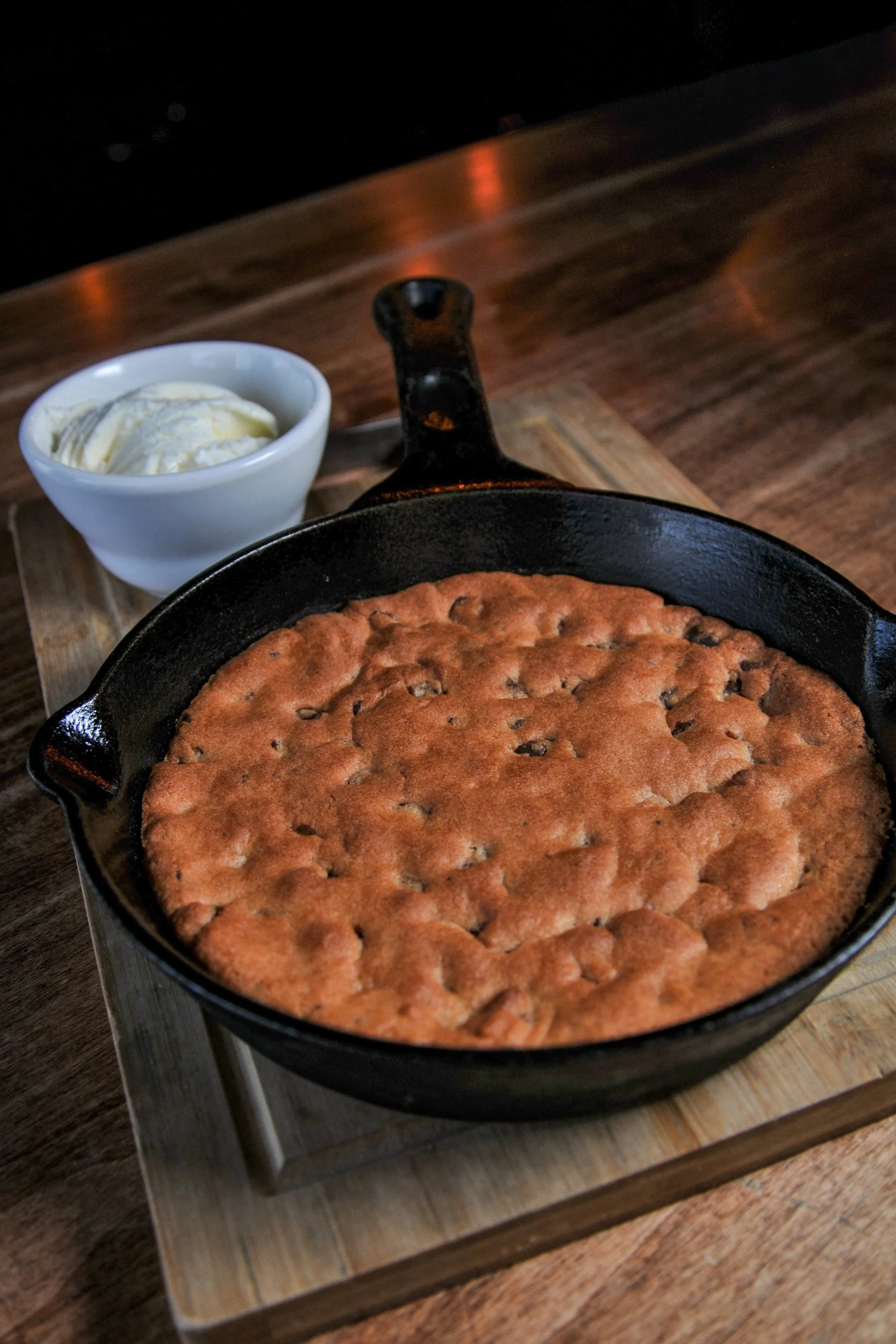 A freshly baked skillet chocolate chip cookie with a side of ice cream in a small white bowl, served on a wooden board on a dark wooden table.