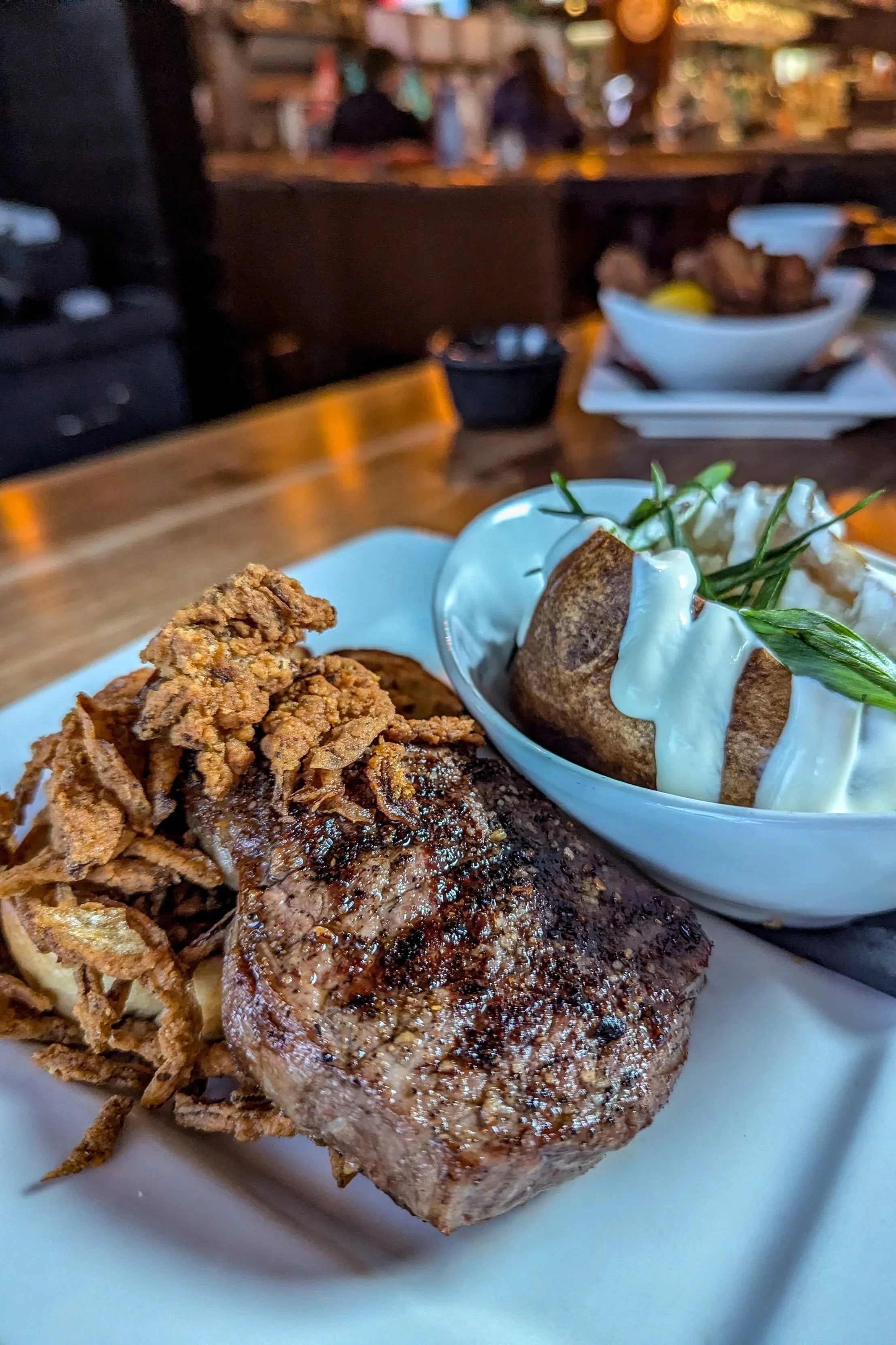 Close-up of a cooked steak with crispy fried onions, served with baked potato topped with sour cream and chives, in a restaurant setting.