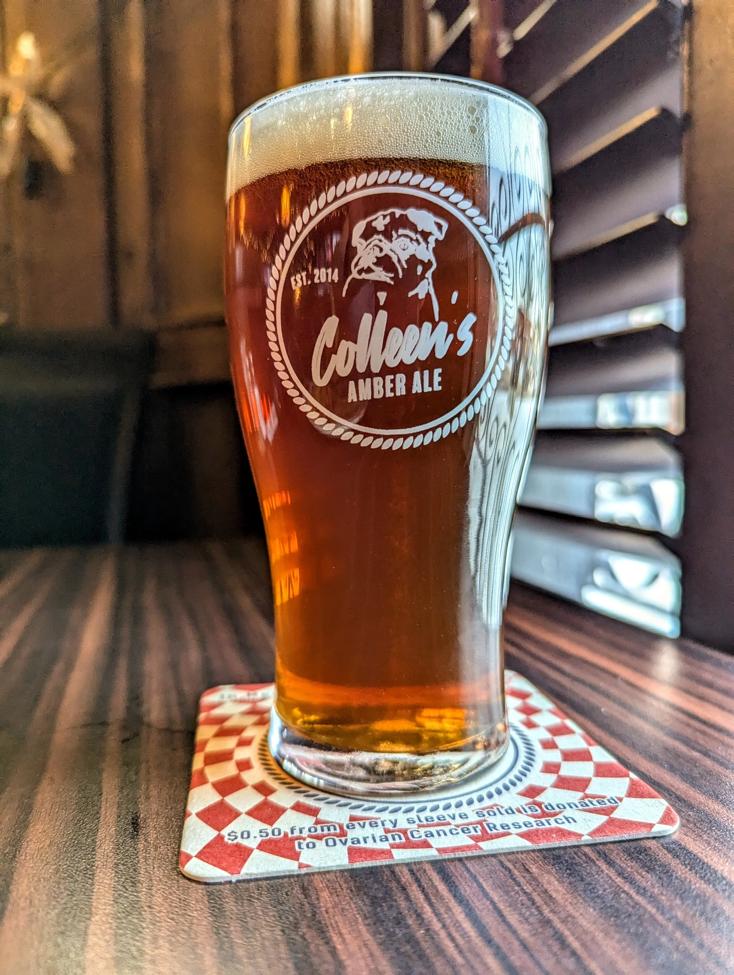 A tall glass of amber beer with frothy head on a wooden table, branded with Colliem's Amber Ale logo, sitting on a first aid-themed coaster that promotes a cancer research donation.