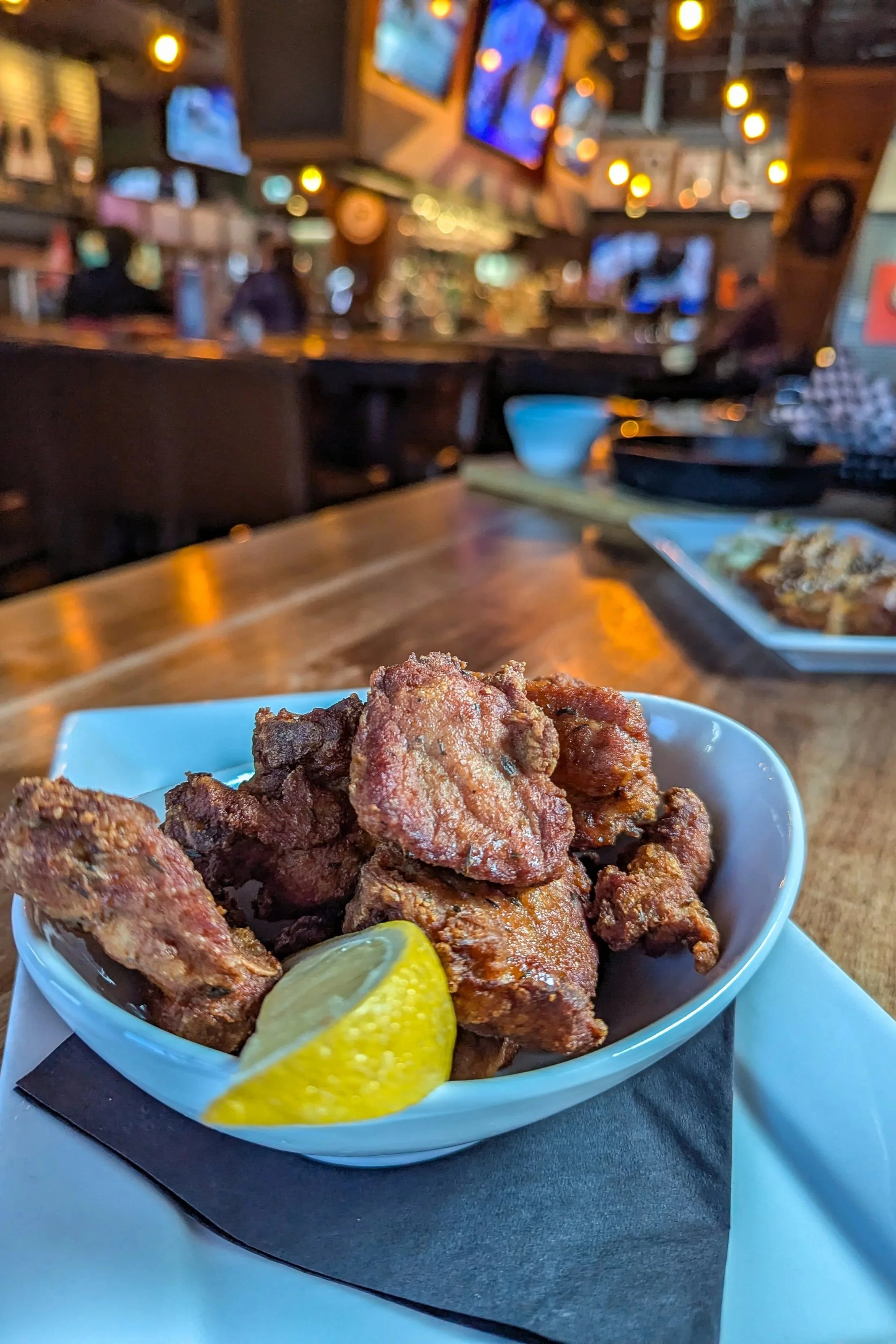 David's Dry Ribs with lemon wedge on a white plate in a restaurant setting with TVs and warm lighting in the background.