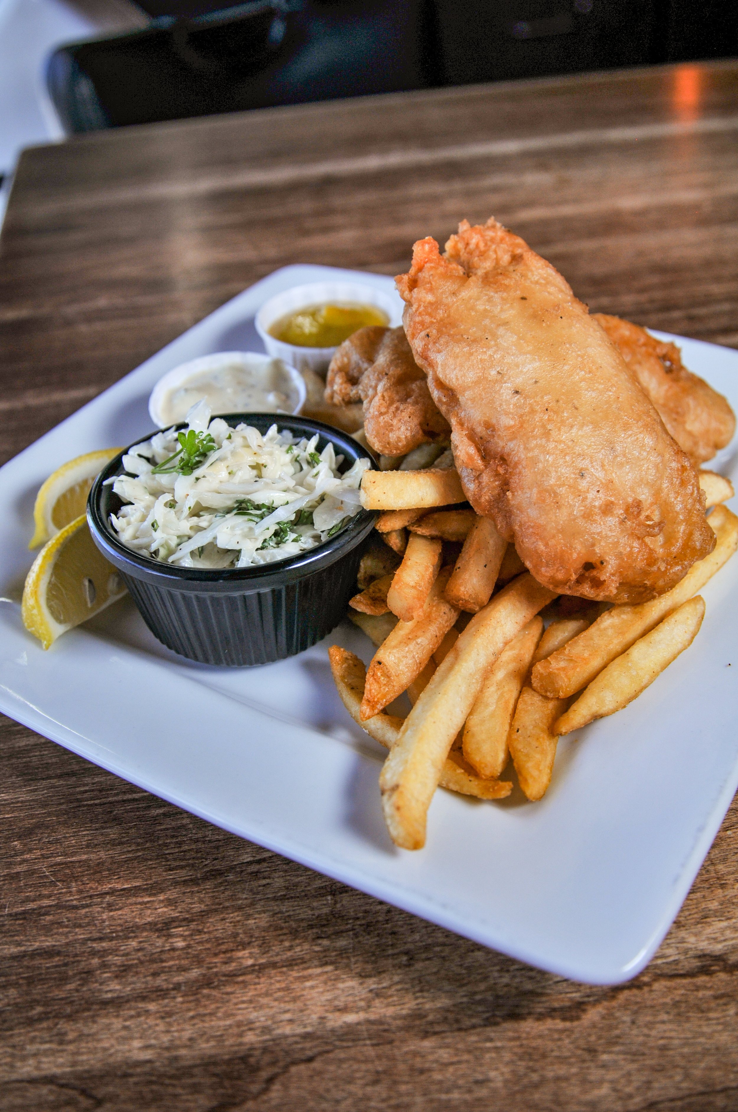 Fish and Chips. Housemade batter, coleslaw, lemon wedges, and dipping sauces on a white square plate.
