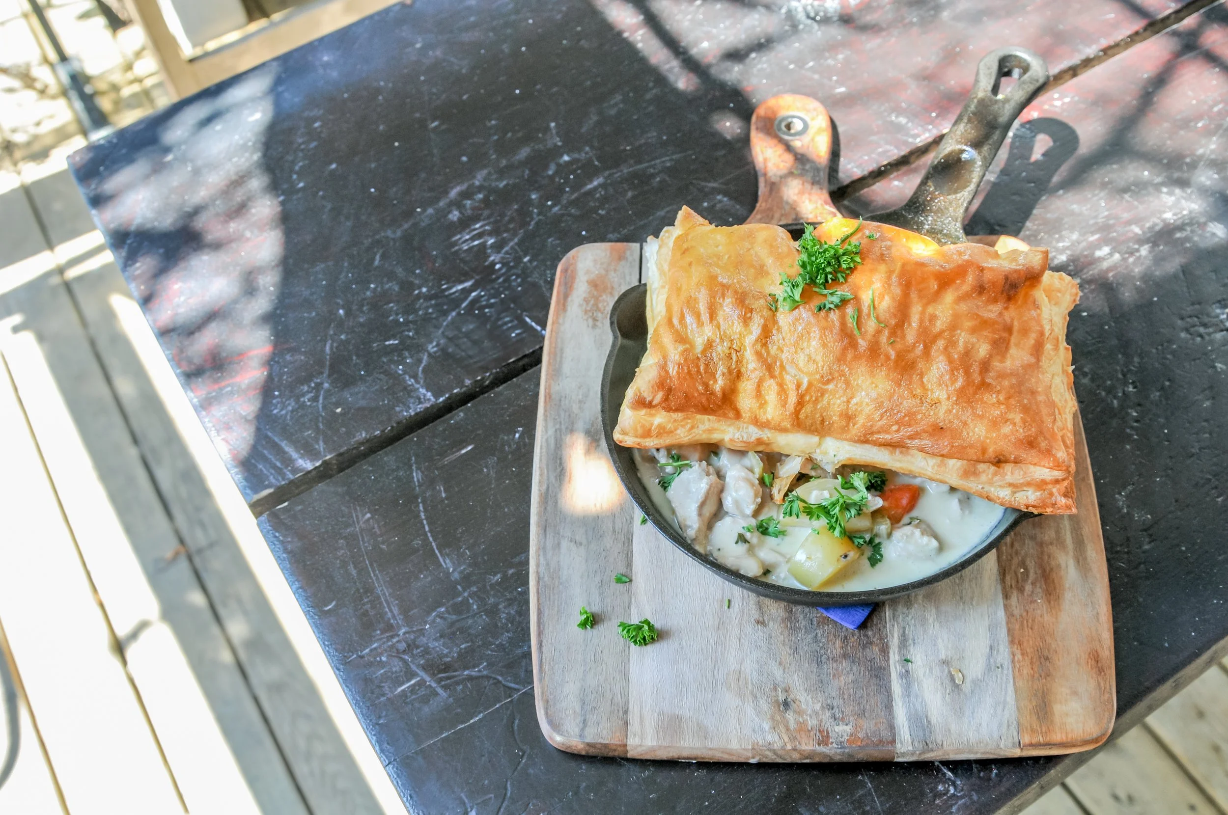 Hearty chicken pot pie with golden puff pastry. served on a black skillet, garnished with fresh parsley, placed on a rustic wooden board on a dark outdoor table.