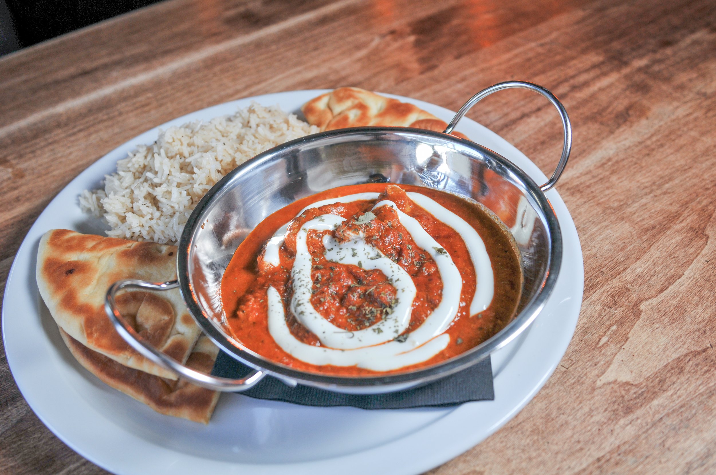 Plate of butter chicken with rice, naan bread, and a curry with cream drizzle and herbs.