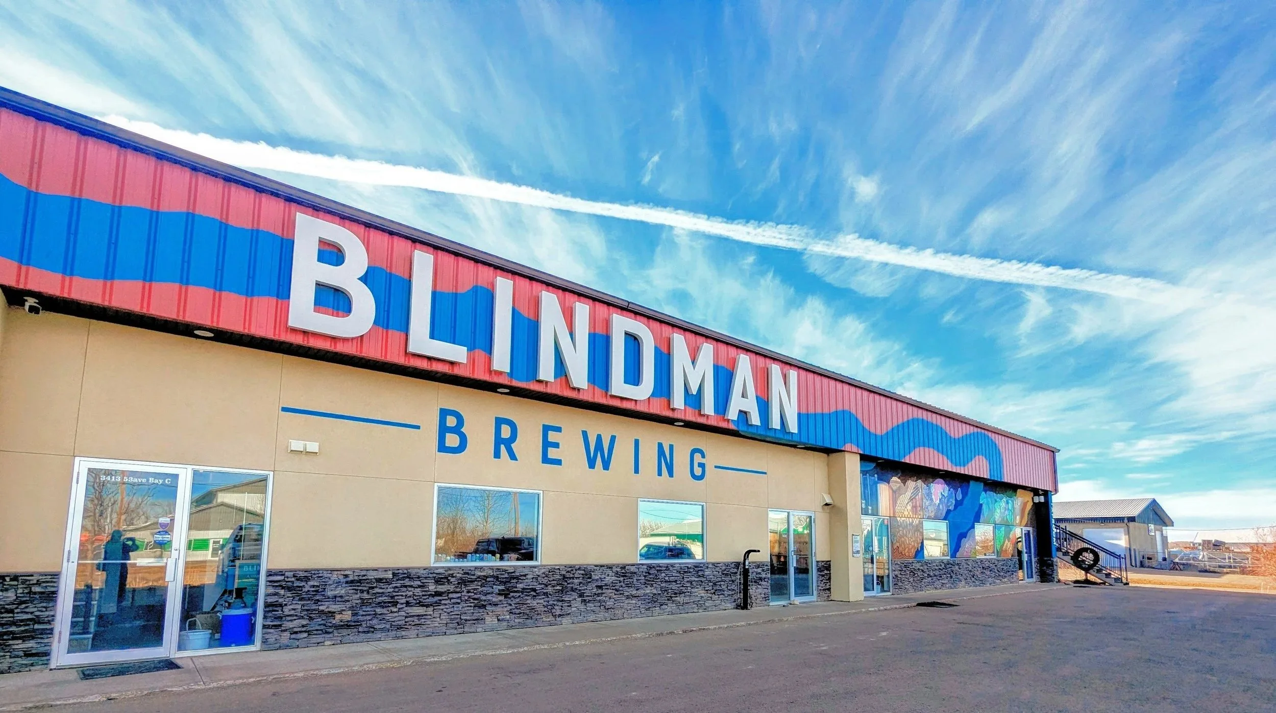 Exterior of a building with a red and blue sign reading 'BLINDMAN BREWING' above the entrance, with a clear blue sky and clouds.