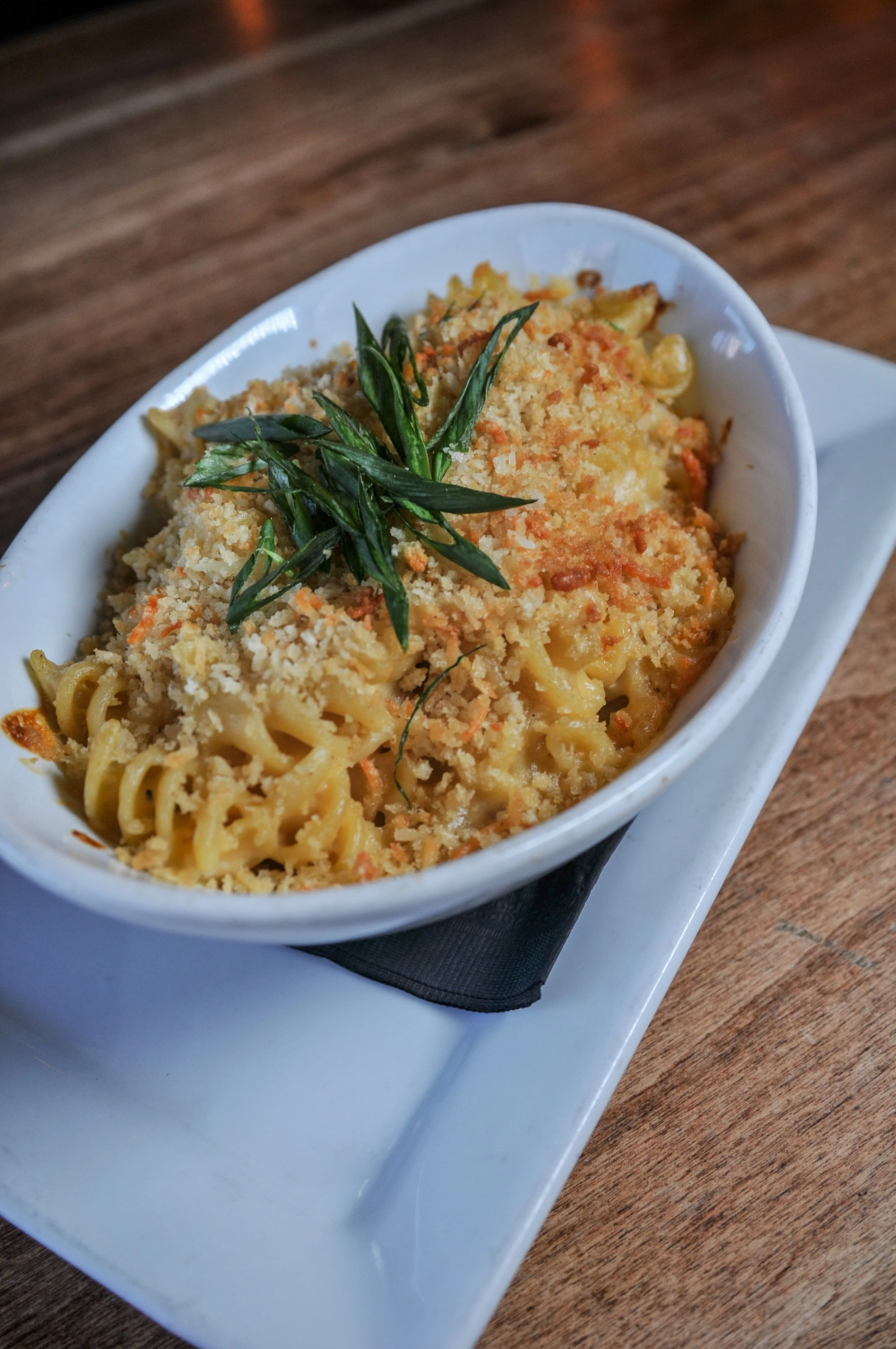 Baked mac n cheese dish with breadcrumbs and green herbs on top, served in a white oval bowl on a white plate with a black napkin, on a wooden table.