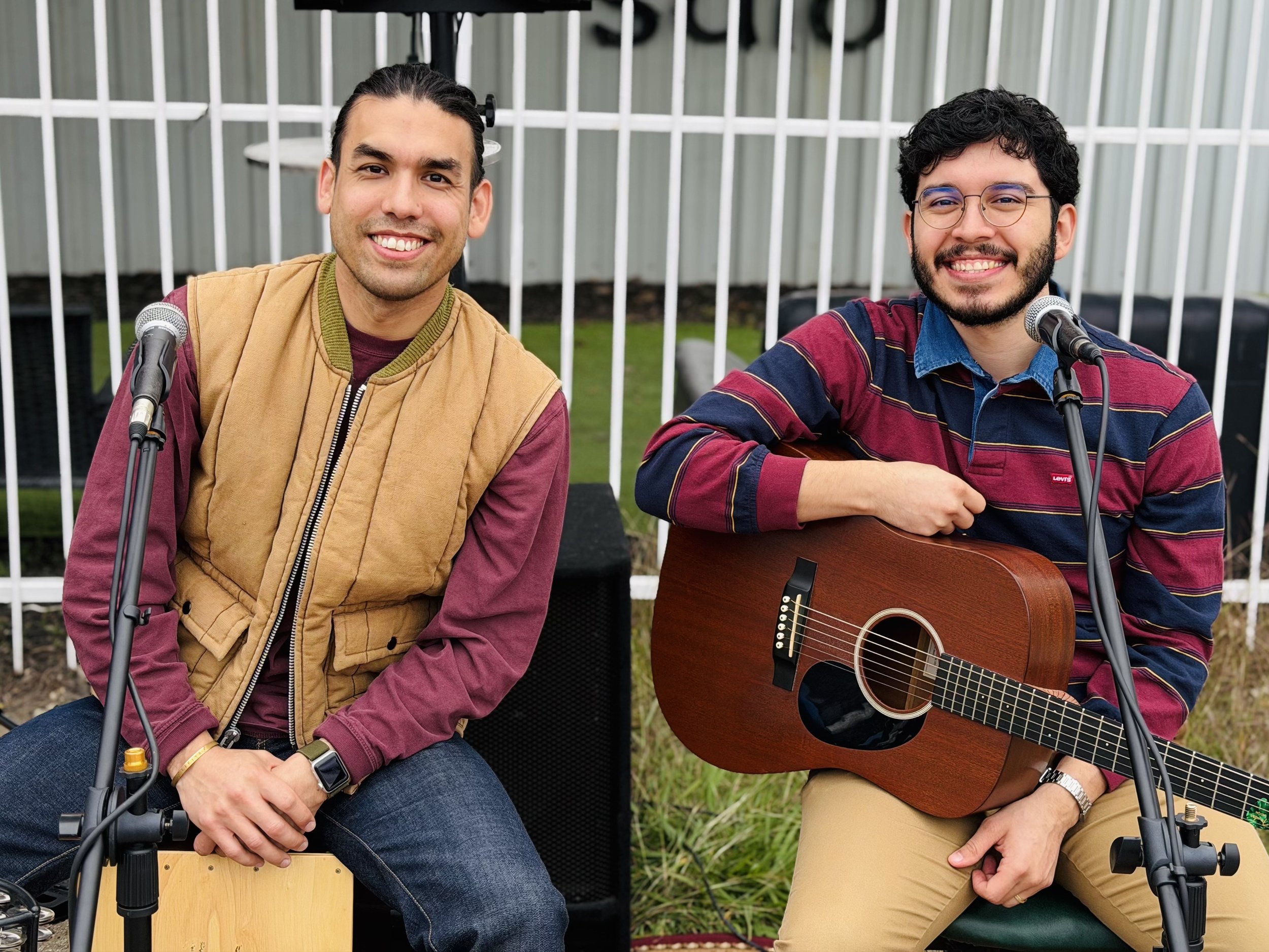 Two musicians seated outdoors in front of microphones, smiling toward the camera; one is playing an acoustic guitar while the other sits on a cajón drum, with speakers and a fence visible in the background
