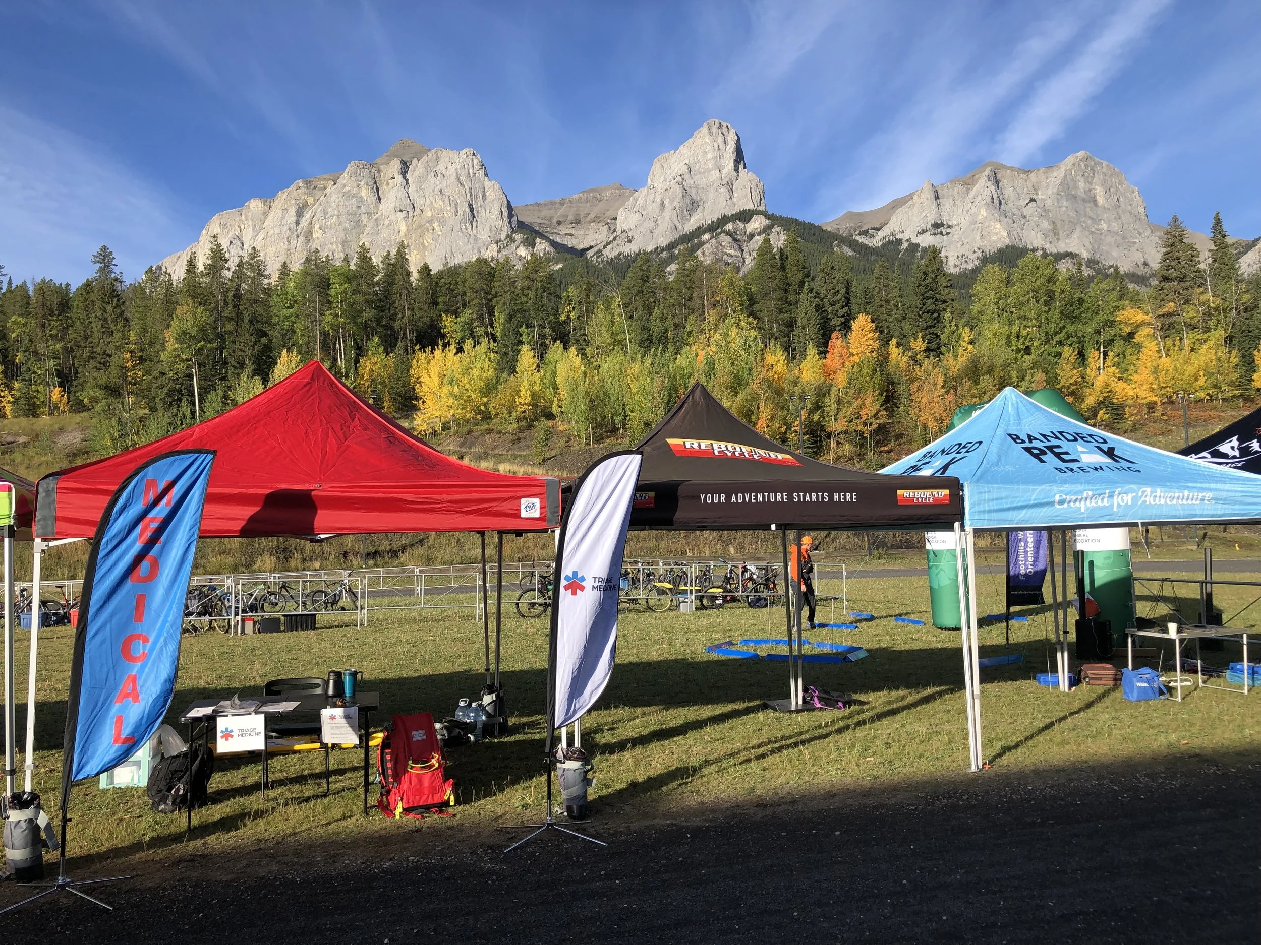 Outdoor event setup with tents, banners, and equipment on grassy field with mountains and trees in the background under a blue sky.