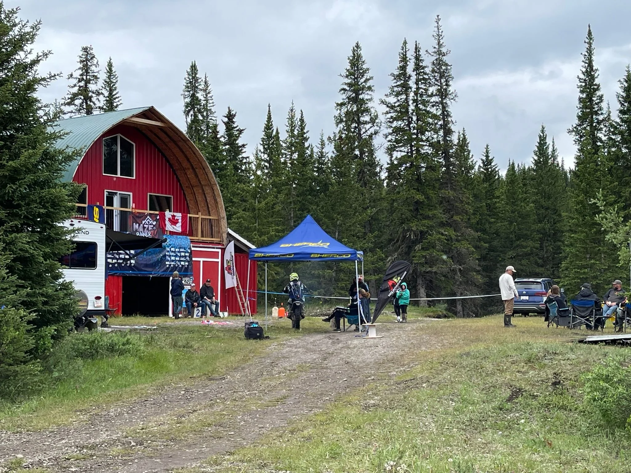 A red barn with a curved roof and trees surrounding it. There are tents set up outside, and several people are gathered, some sitting and some standing, in an outdoor event area.