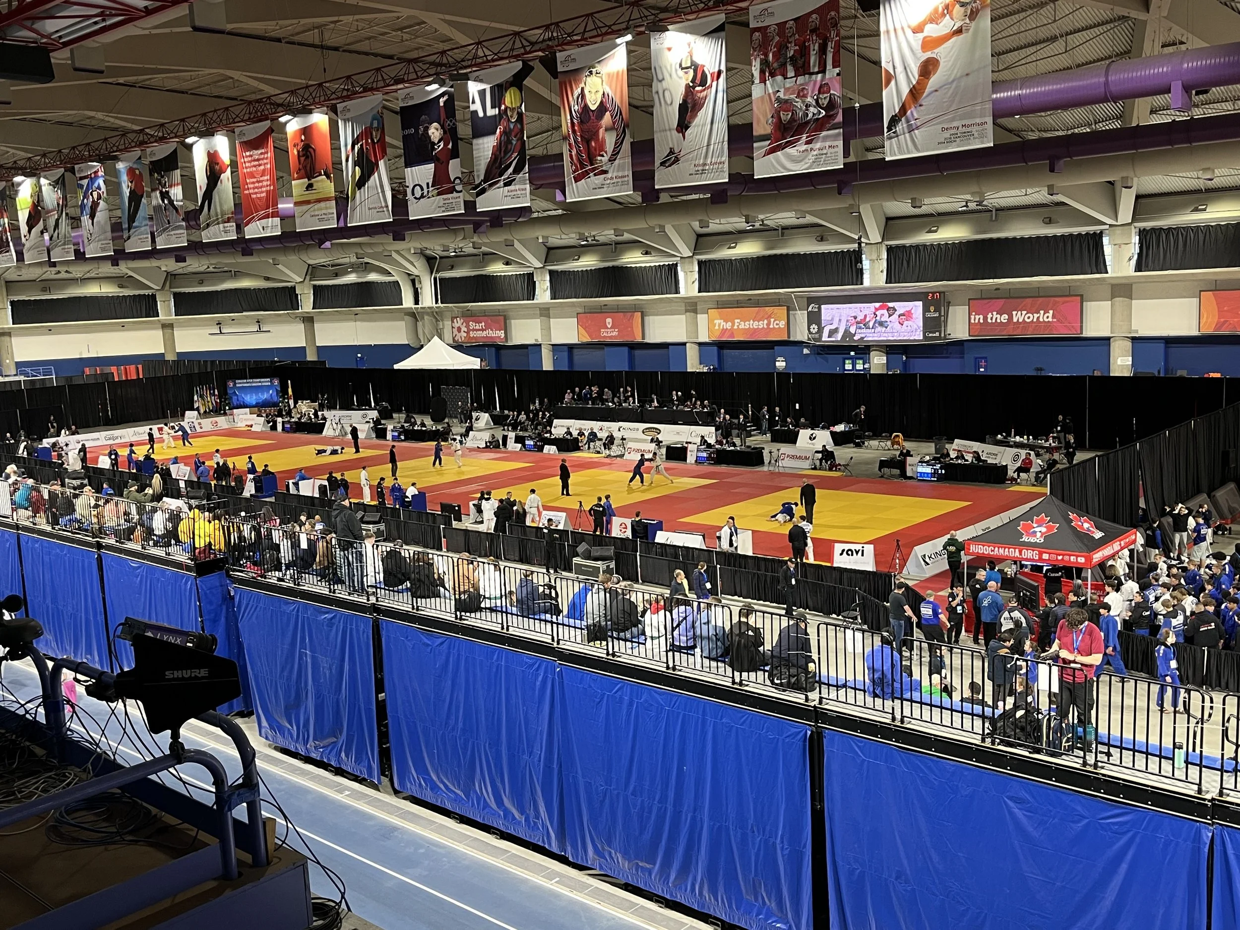 Indoor sports arena with a wrestling or judo event taking place. Spectators seated behind a barrier, athletes on mats, banners hanging from the ceiling, and a digital display board.