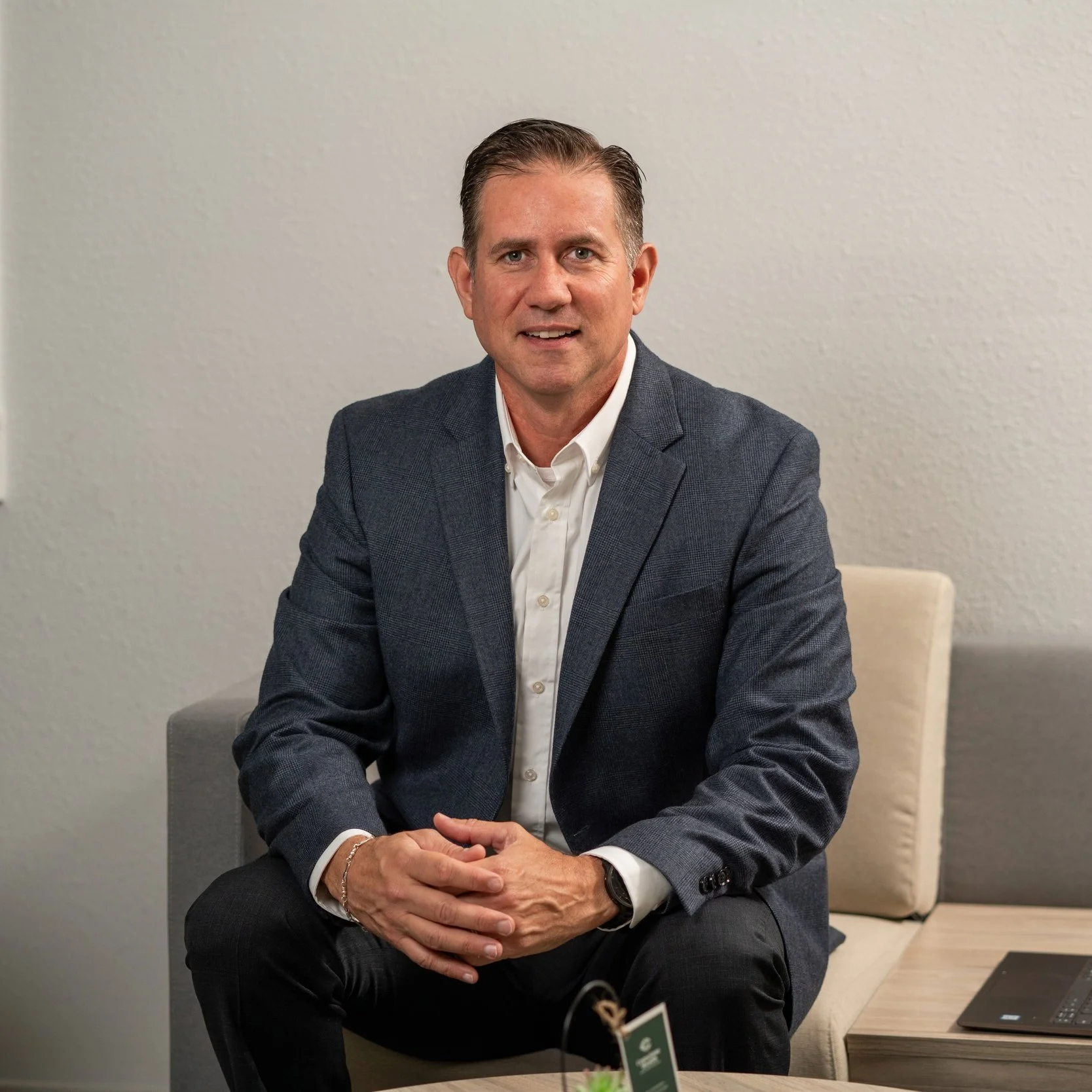 A man in a gray suit jacket and white shirt sitting on a beige and gray bench, with a light-colored wall behind him.