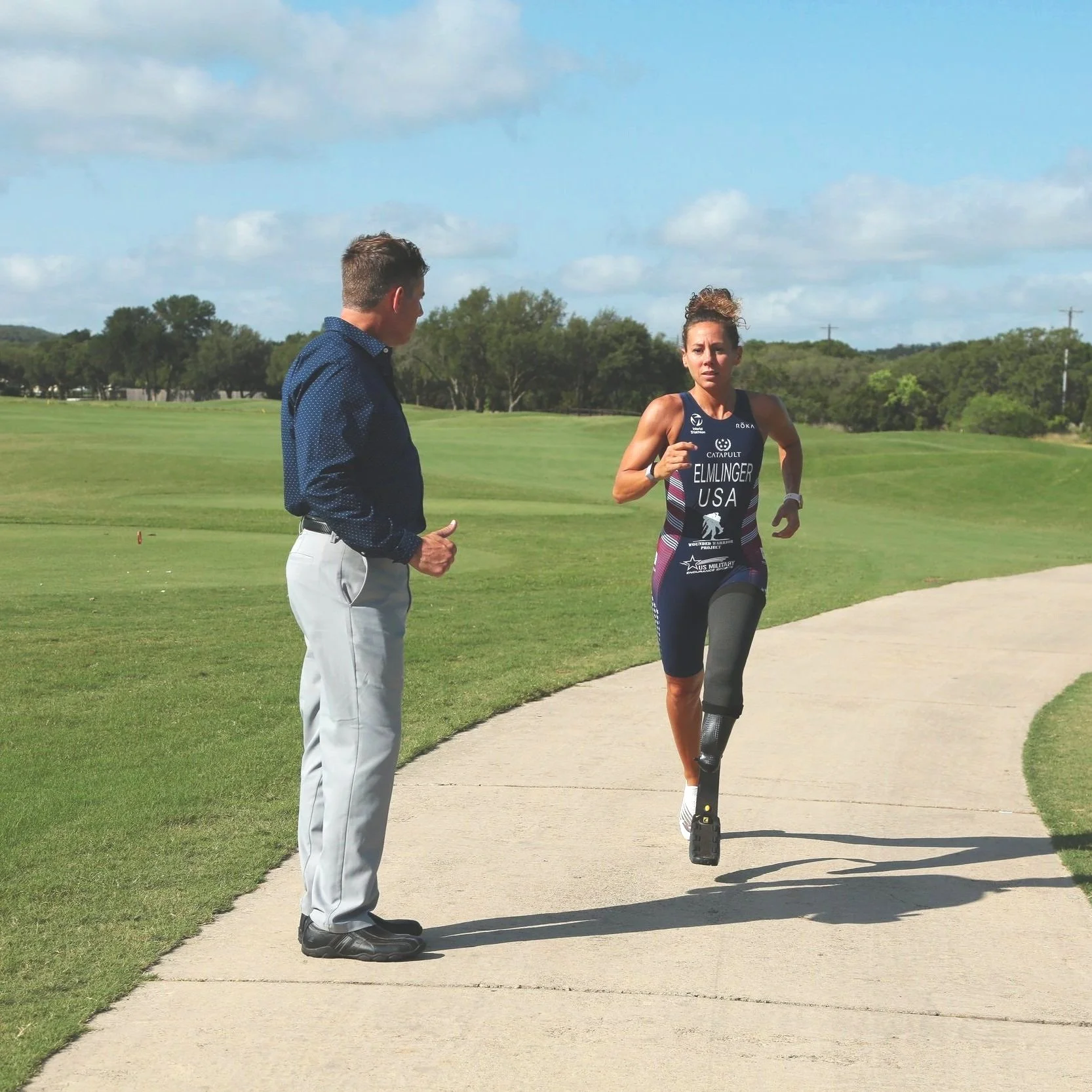 A woman with a prosthetic leg running on a sidewalk next to a man in a city park or golf course, with trees and a cloudy sky in the background.