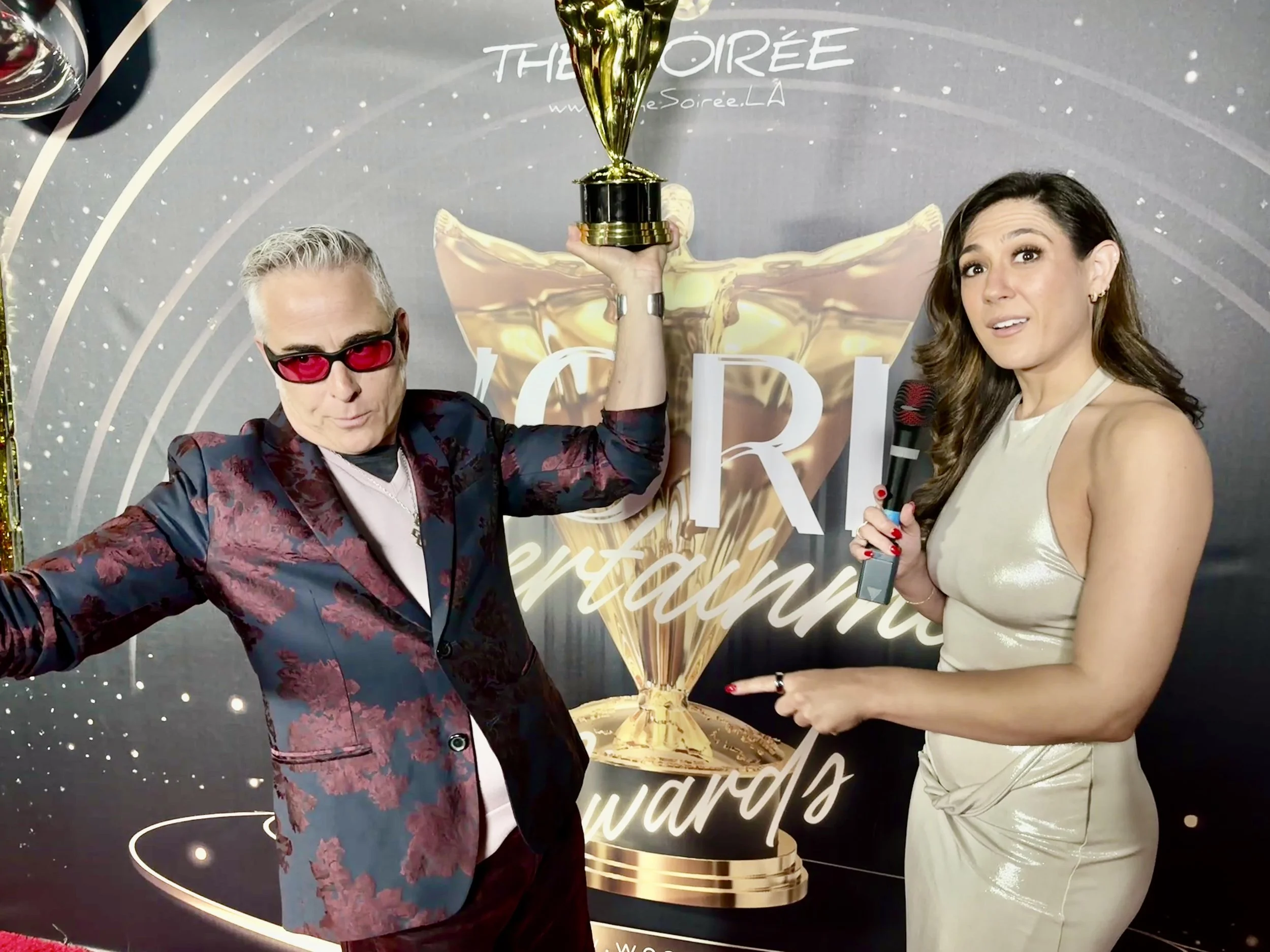 A man wearing red sunglasses and a floral blazer holding a trophy, standing beside a woman in a cream-colored dress holding a microphone, at an award ceremony with a backdrop that reads "The Soirée" and "Awards."