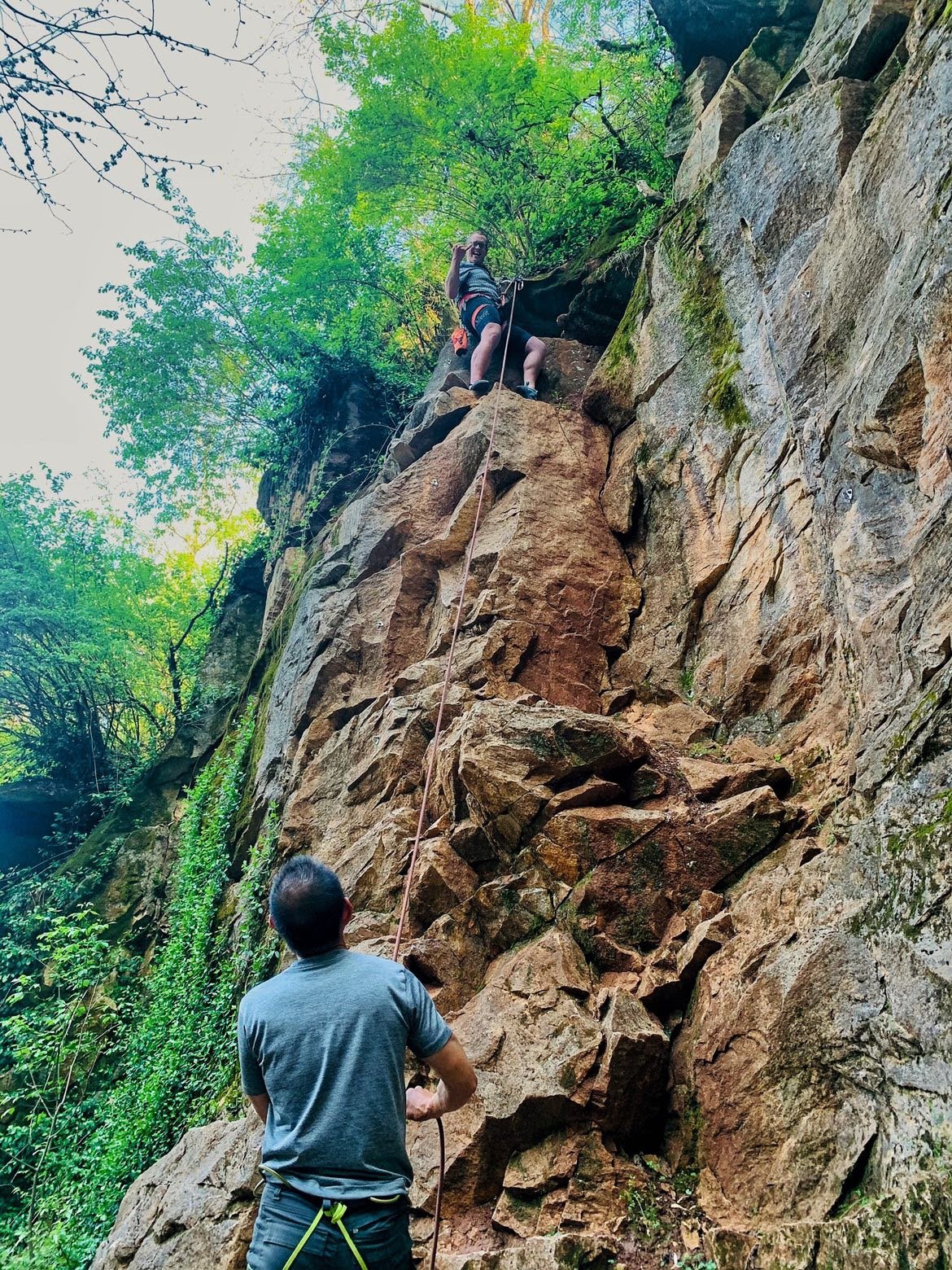 A person belaying a climber on an outdoor rock climbing wall in a forested area.