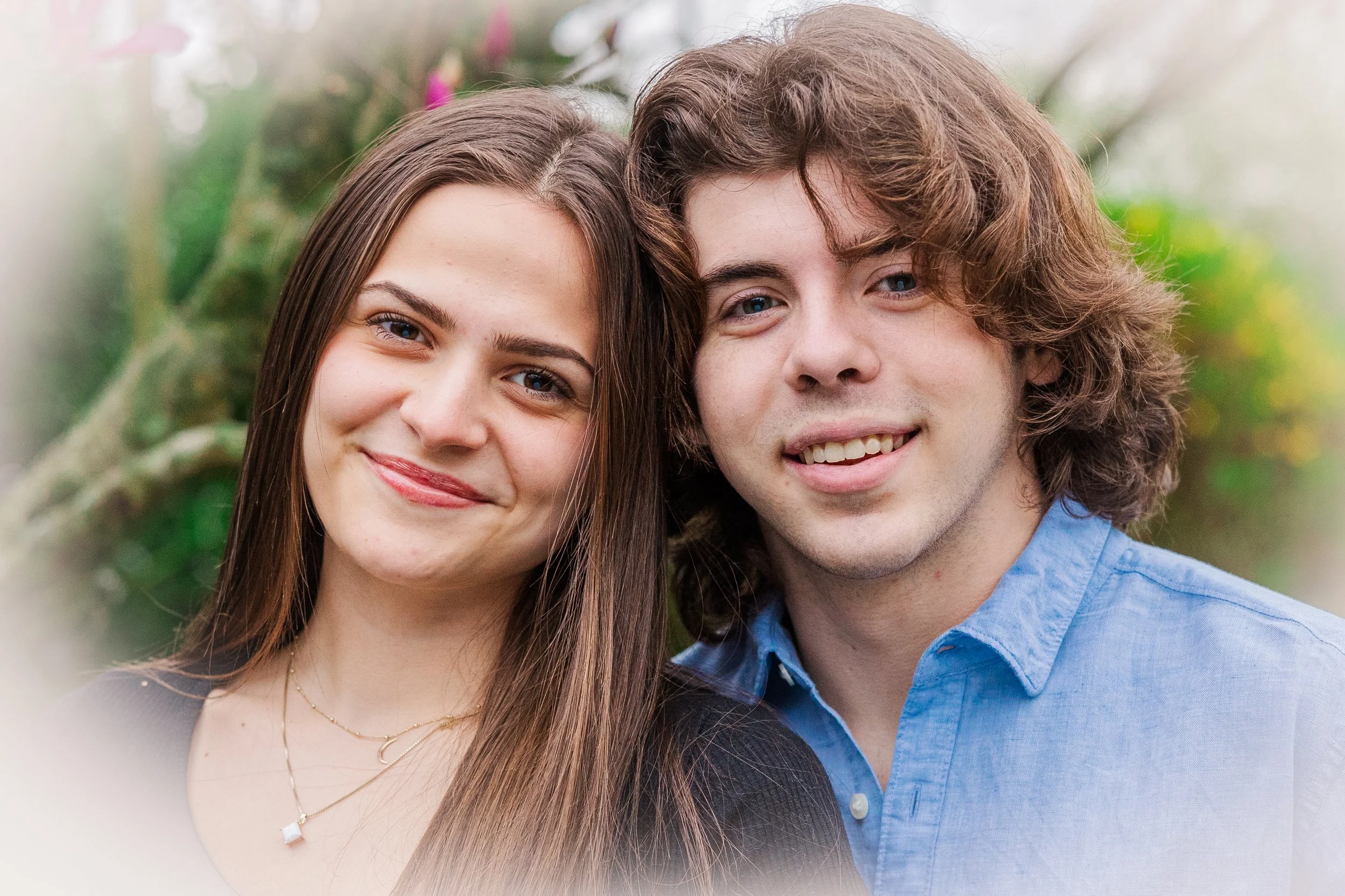 Close-up of a smiling young woman with long brown hair and a young man with curly brown hair, standing outdoors with blurred greenery in the background.
