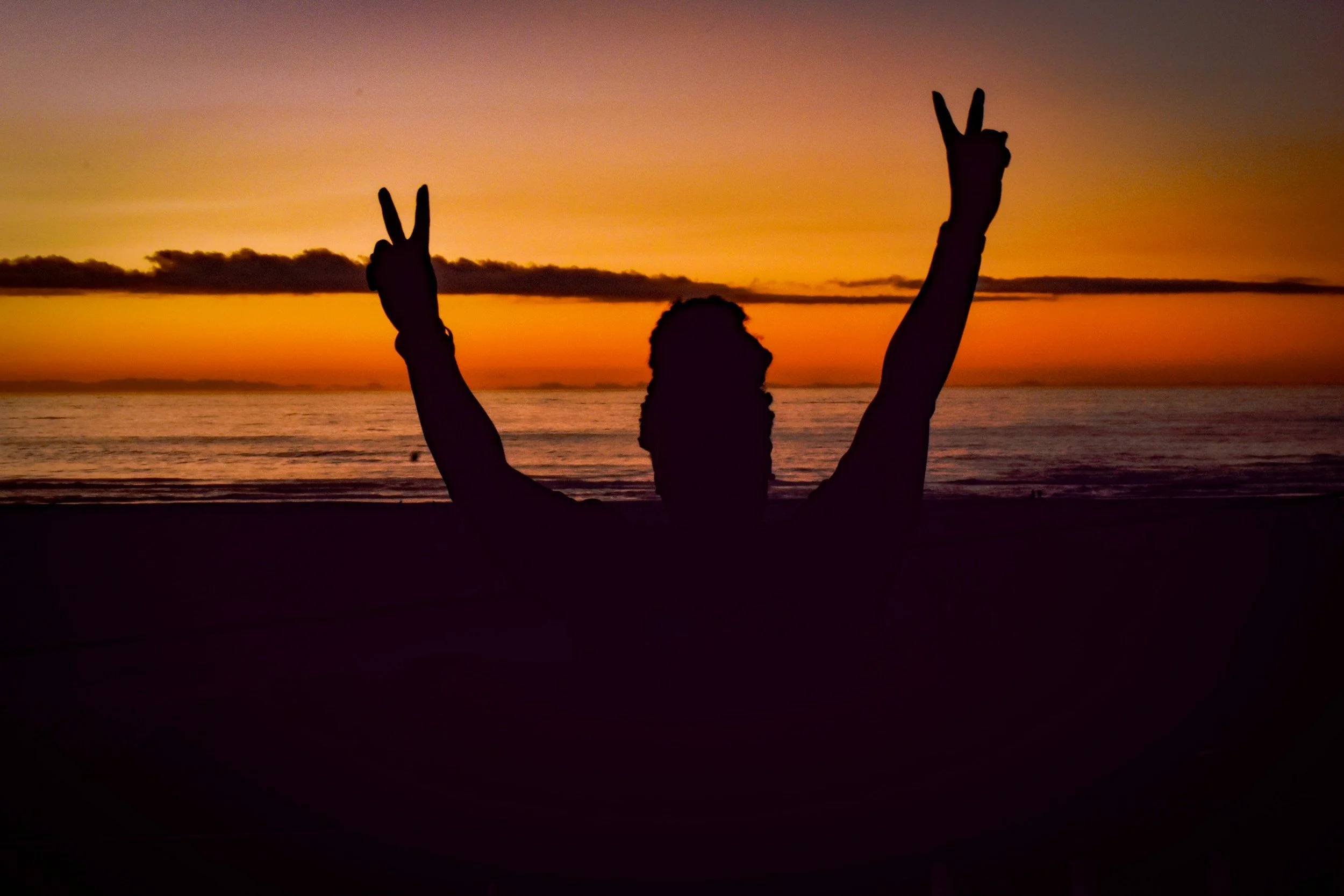 Silhouette of woman near a beach and sunset with arms lift and making the peace sign with both hands.