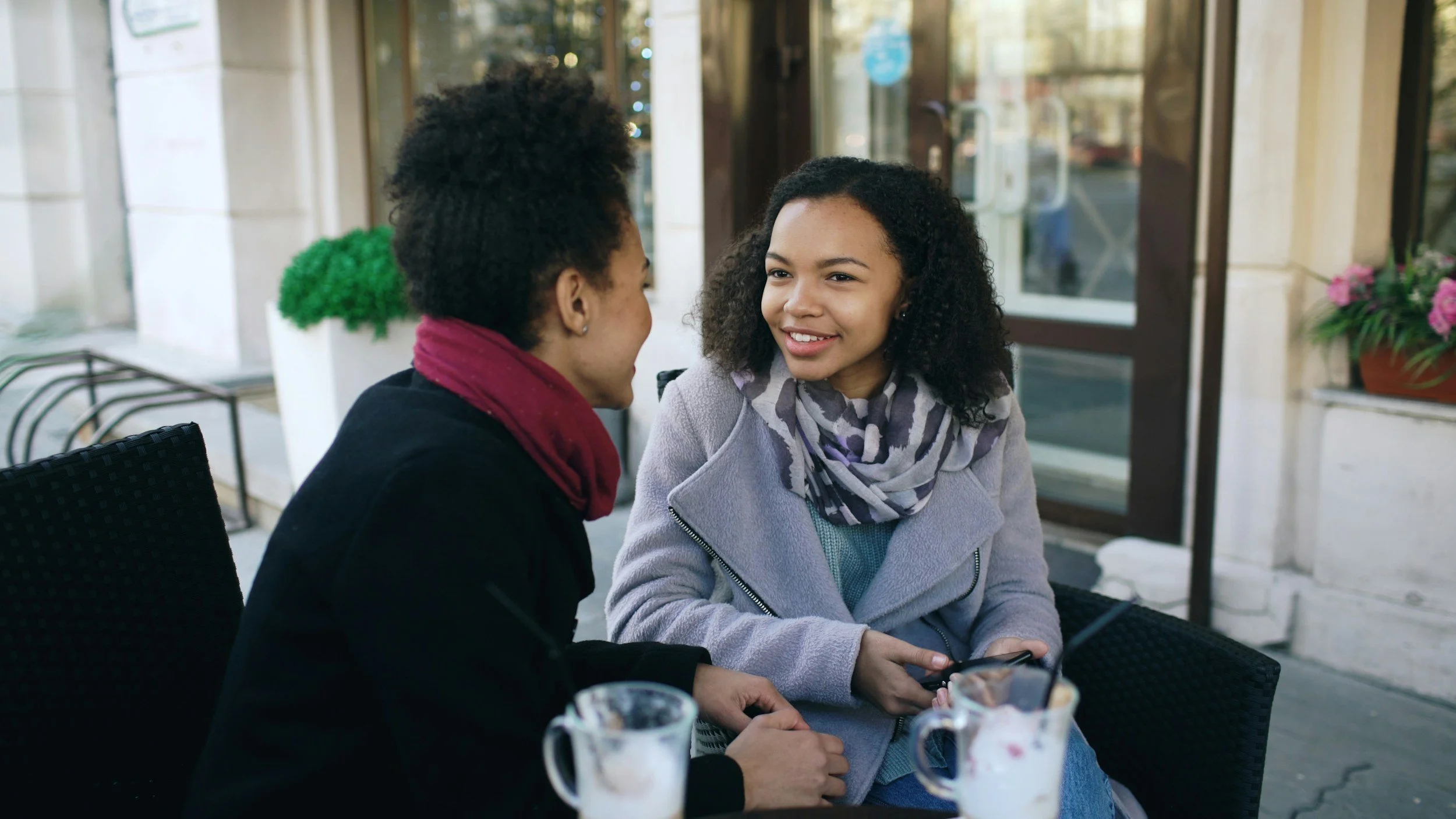Two African American moms sitting outside at a cafe talking with gentle smiles.