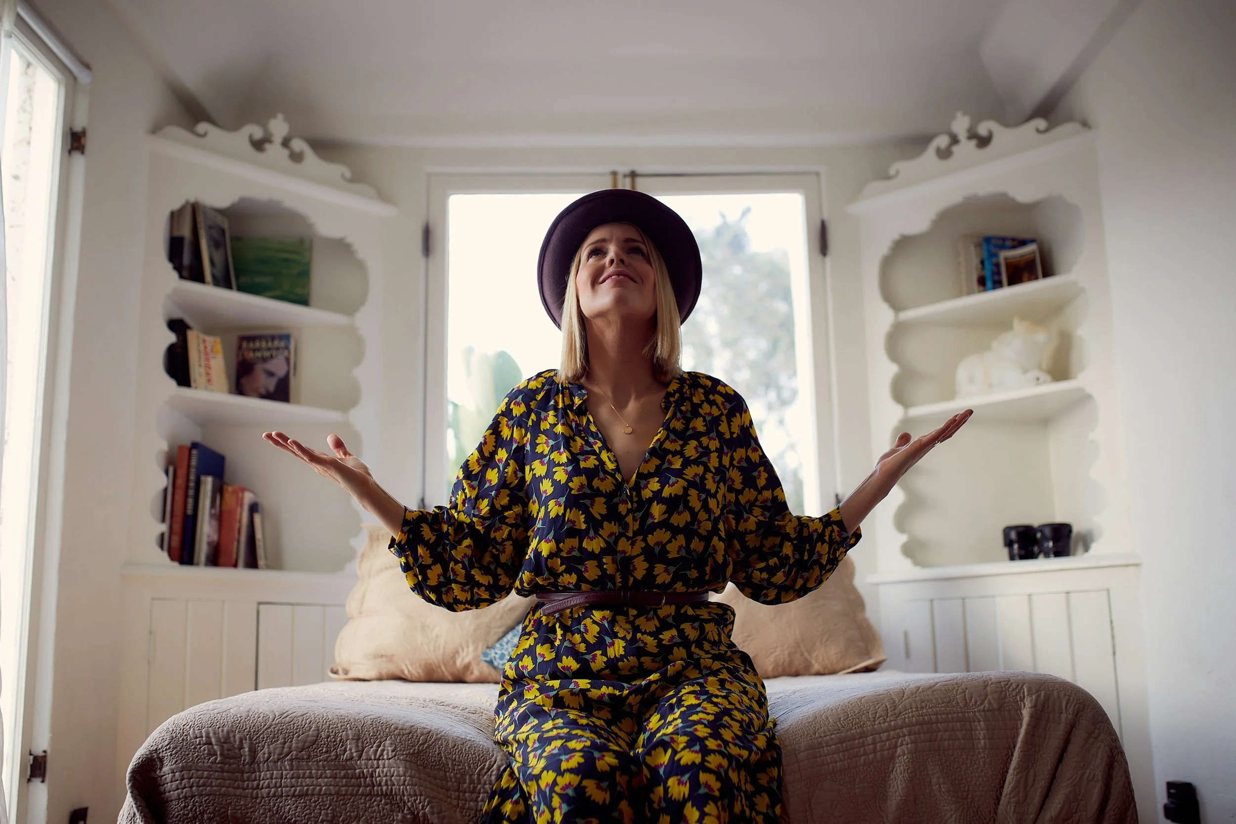 Christian mom wearing a yellow dress sitting the edge of the bed with arms lifted in worship.