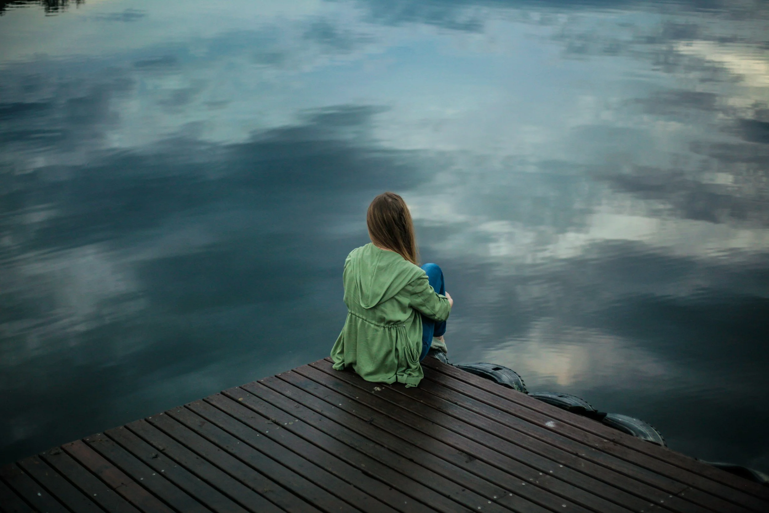A woman with a green jacket sitting on a dock of a lake.