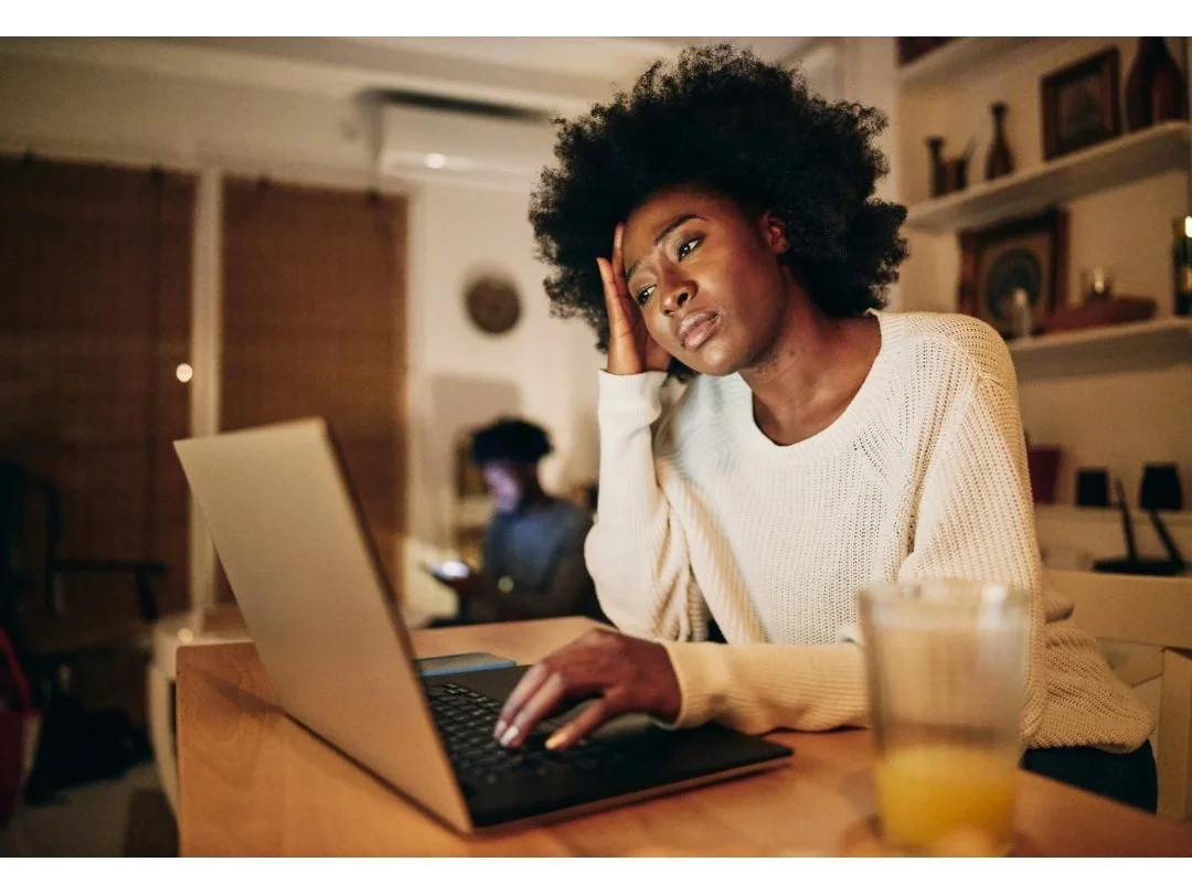 Black mom tired at the computer with teenage son in the background.