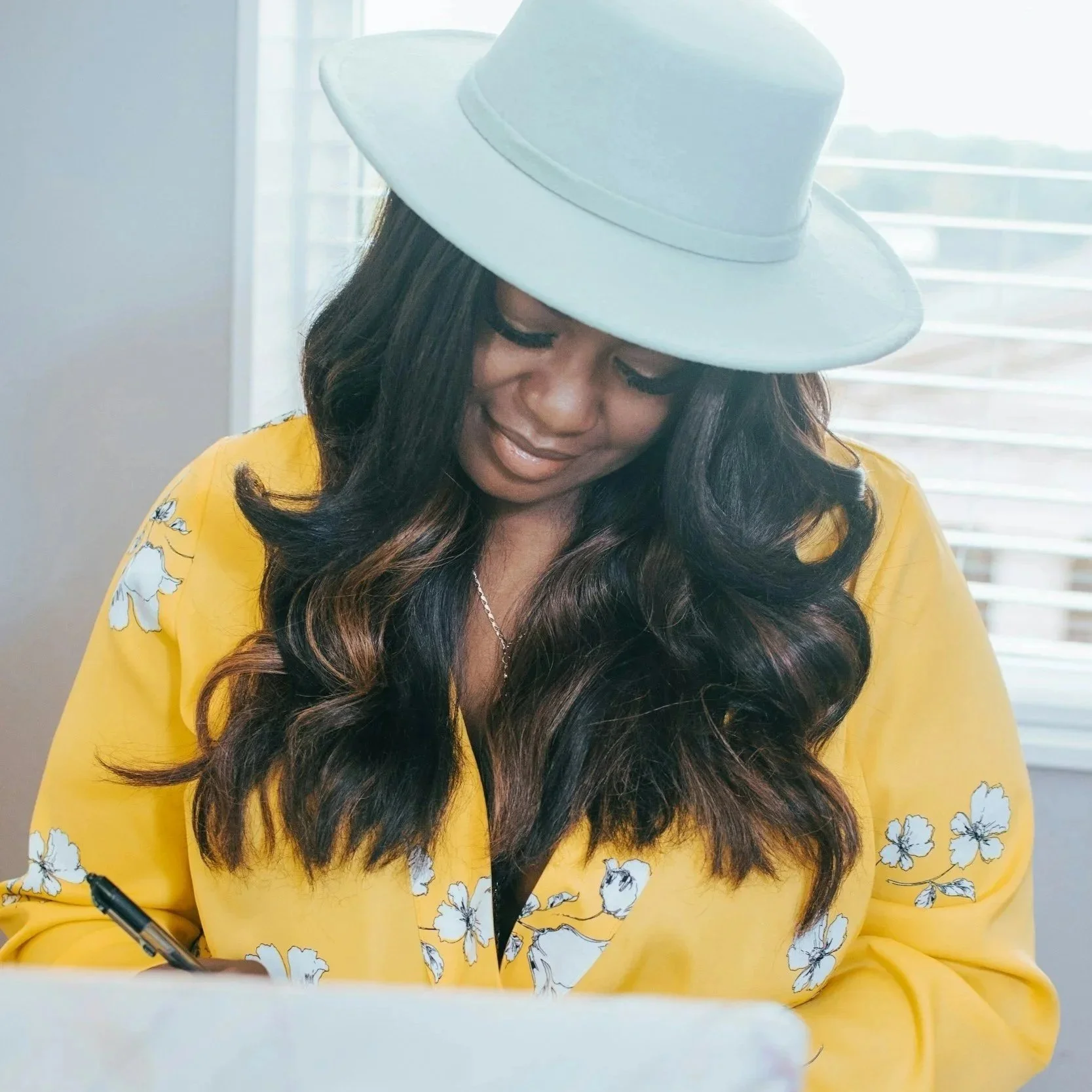 Woman with yellow blouse, and white brim hat smiling and working at her desk.