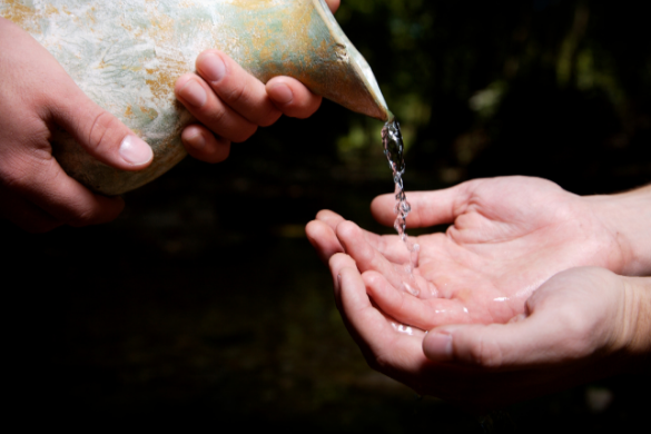 Someone pouring water from a pitcher onto the hands of another person's hands.